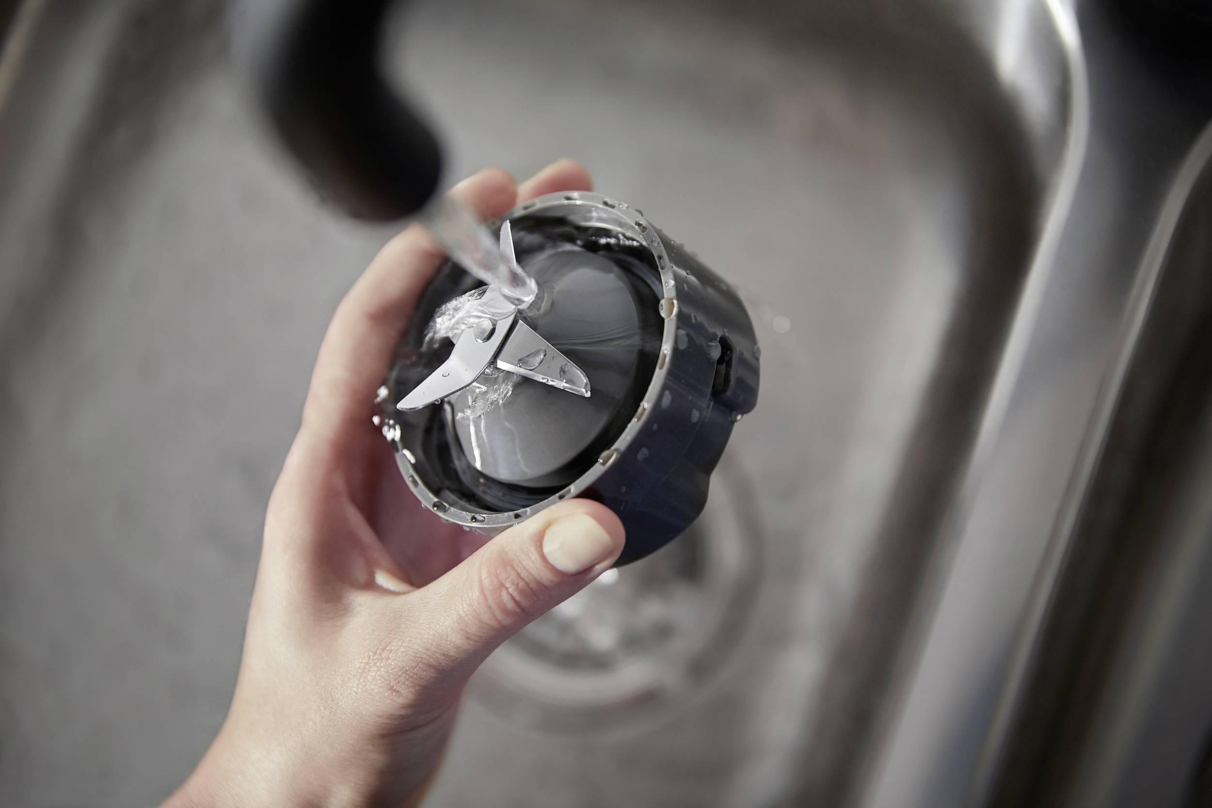 A hand is holding a mixer blade under running water in a sink to clean it.
