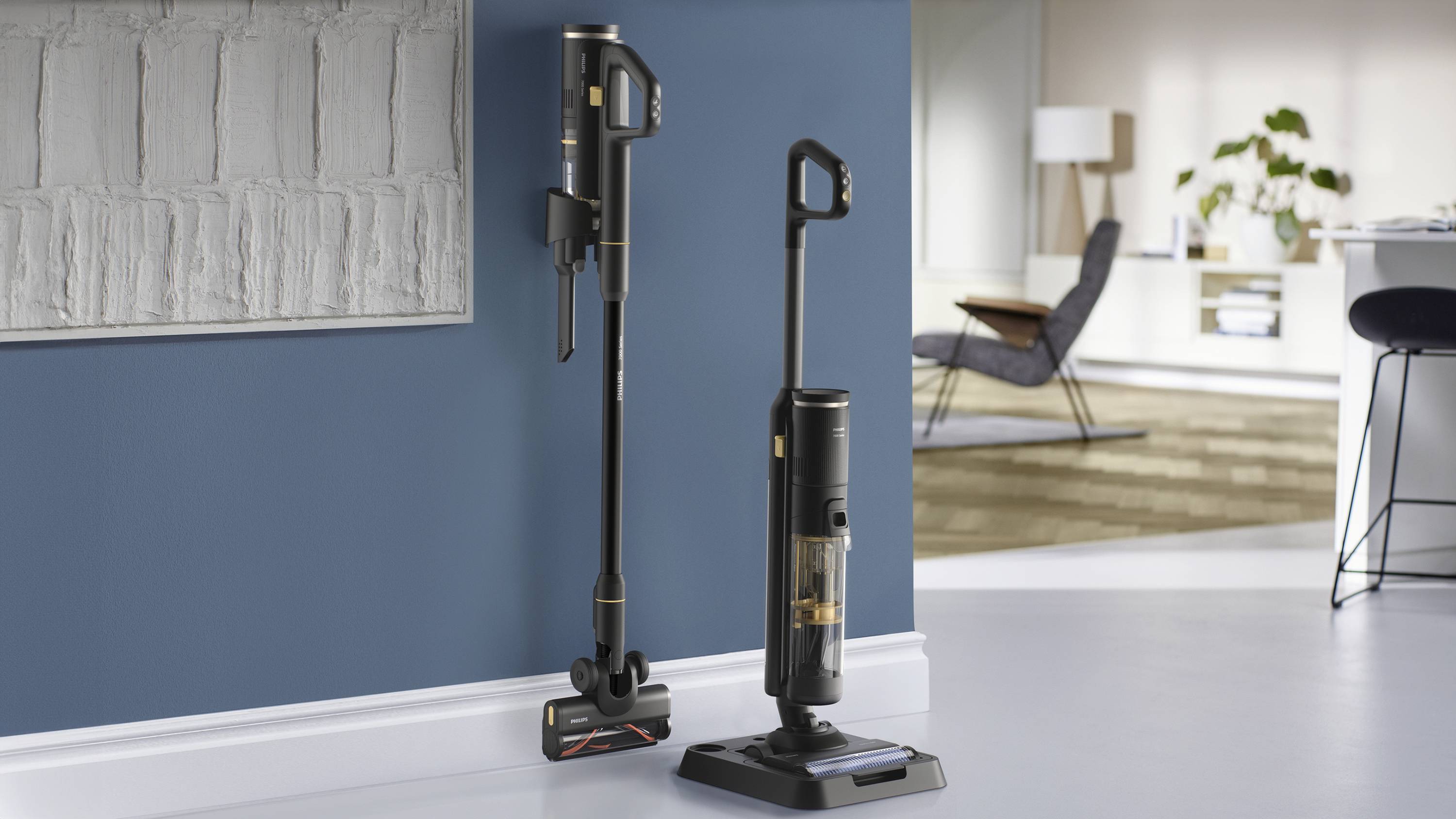 Cordless vacuum cleaners stand against a blue wall in a modern living room. In the background, an armchair and a table with a lamp.