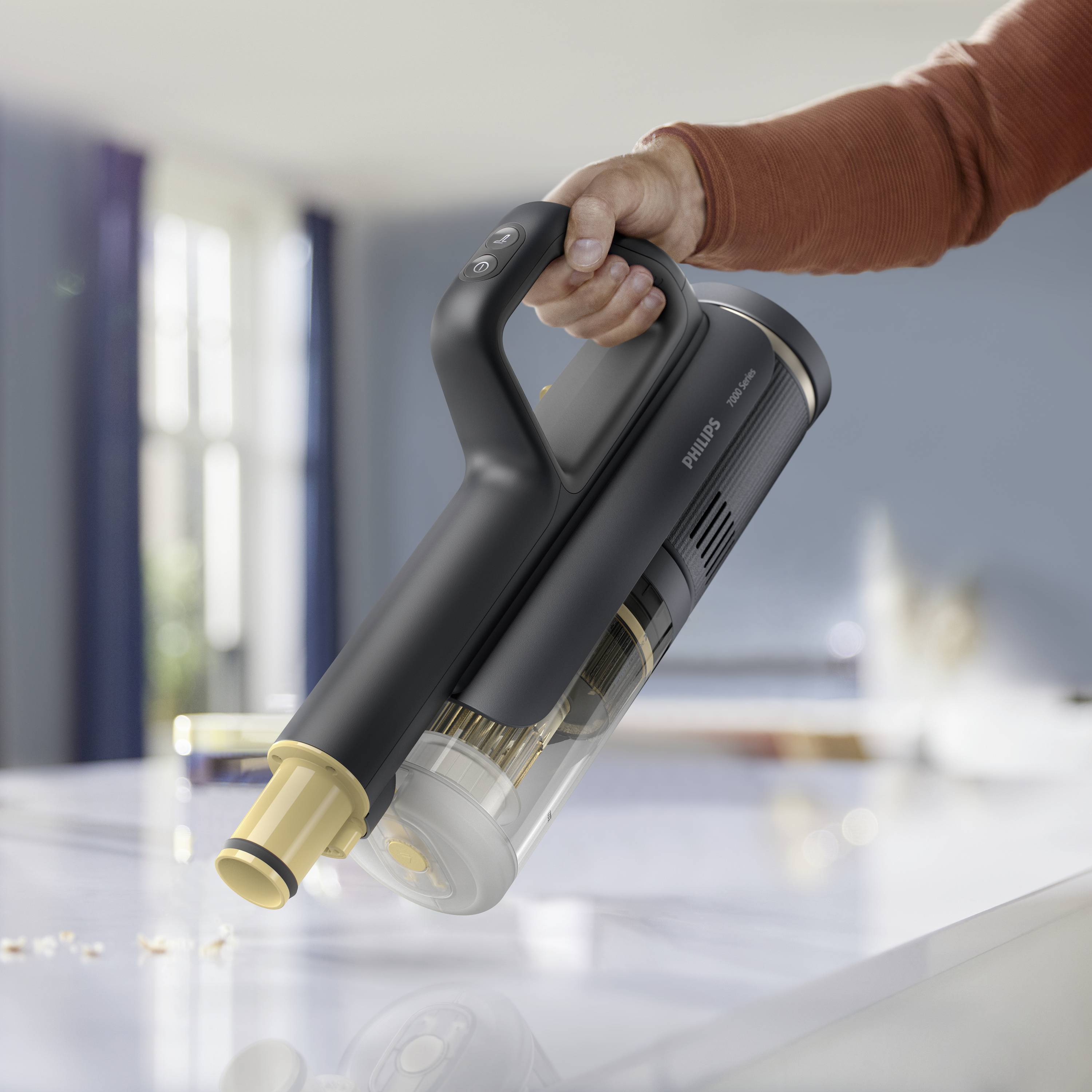 A person is holding a handheld vacuum cleaner, which is sucking up crumbs from a table. A bright room can be seen in the background.