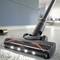 A vacuum cleaner with an illuminated nozzle is cleaning a floor. A blurred room can be seen in the background.
