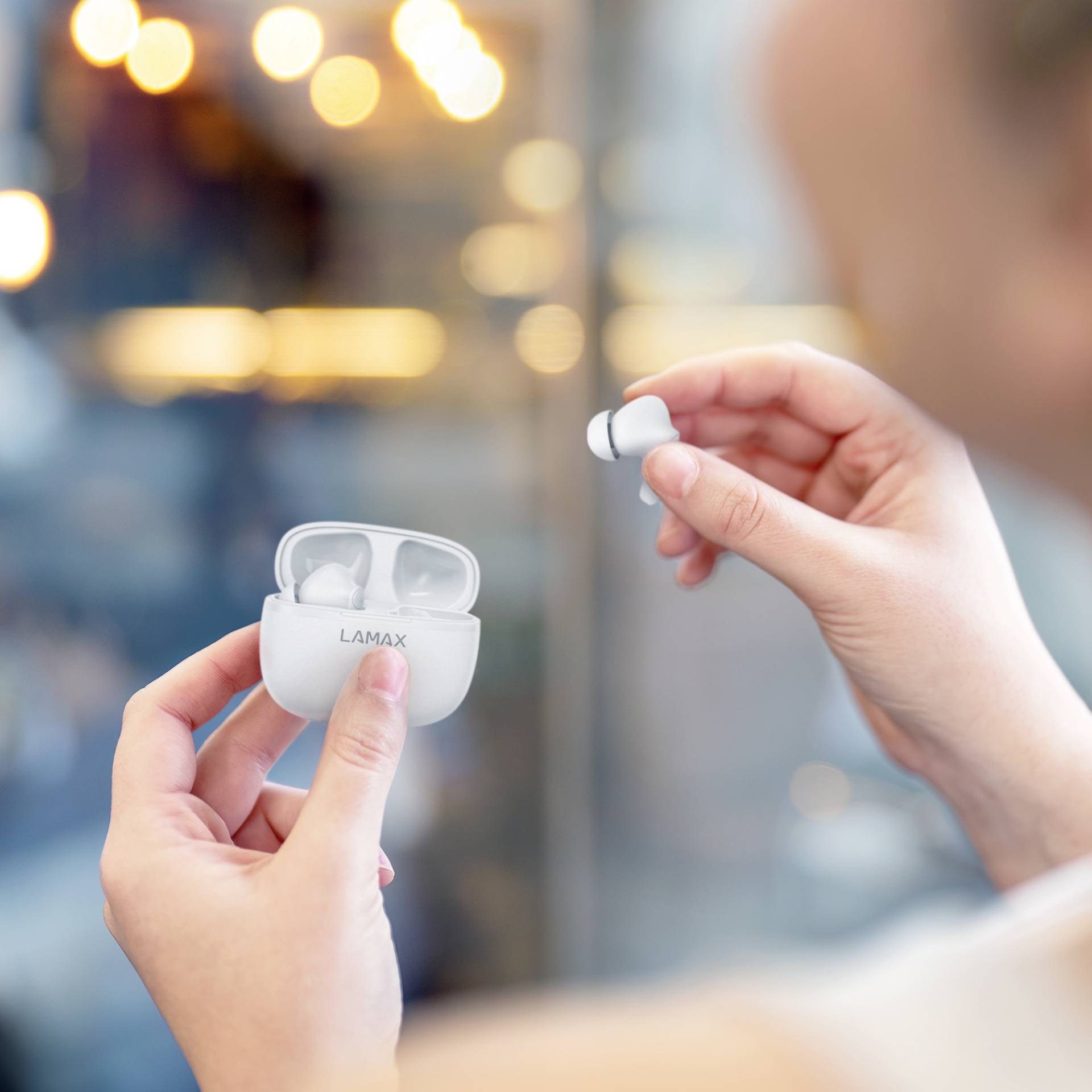A person is holding wireless earbuds in their hand while opening the charging case. Background blurred with lights.