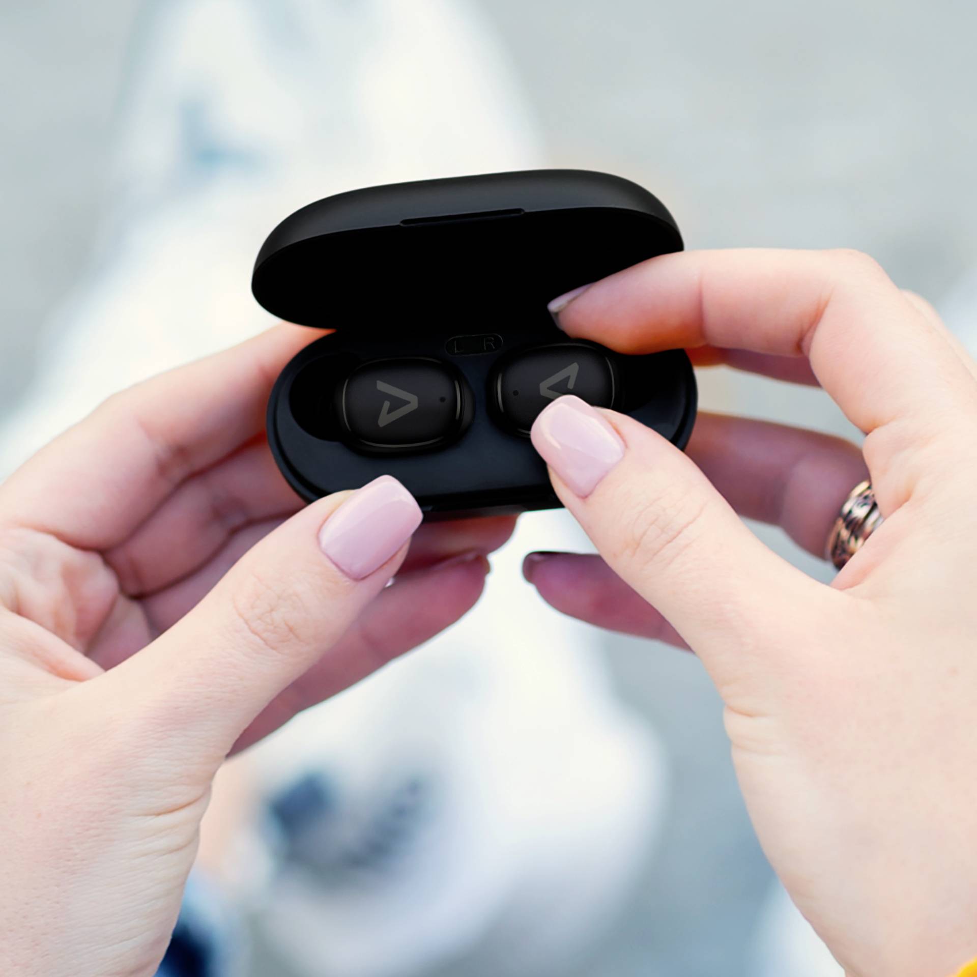 A person is holding an open case with wireless earbuds in their hand, ready to use them. They are outside.