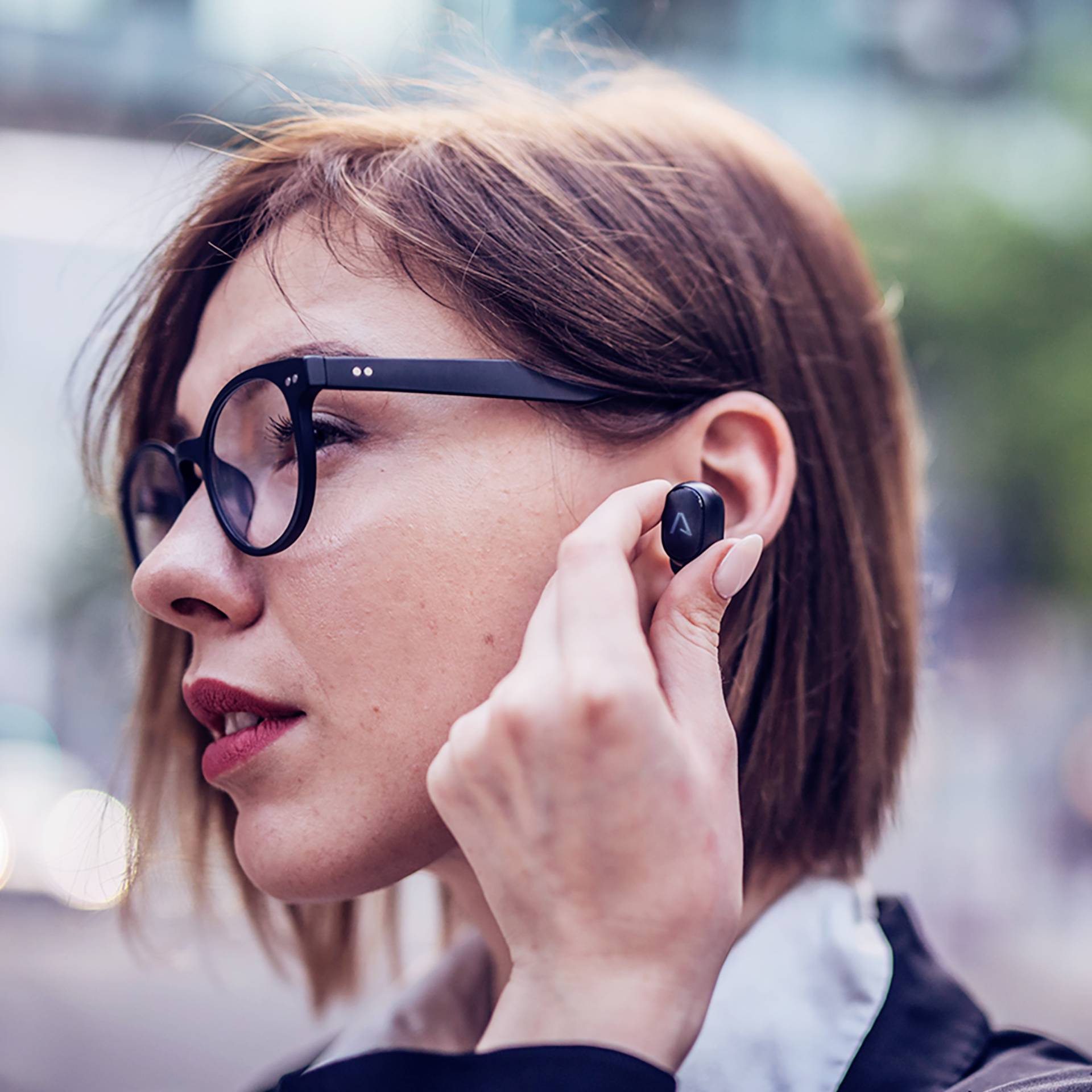 A person wearing glasses is inserting an earphone into their ear. They are wearing a black top and are outside.