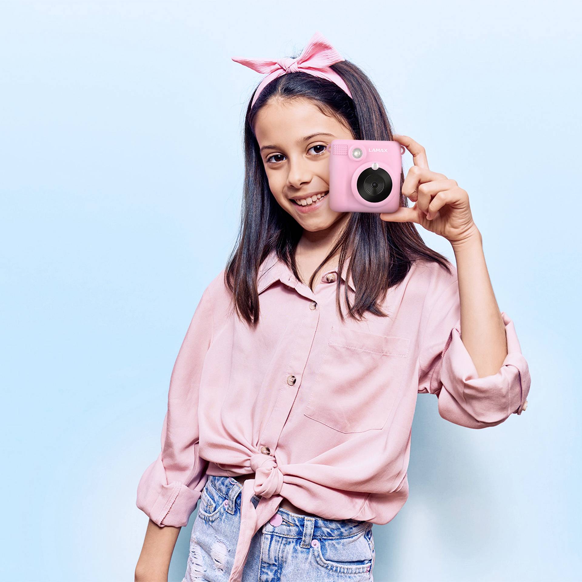 A smiling girl wearing a pink hairband and shirt is holding a pink camera, standing in front of a light blue background.