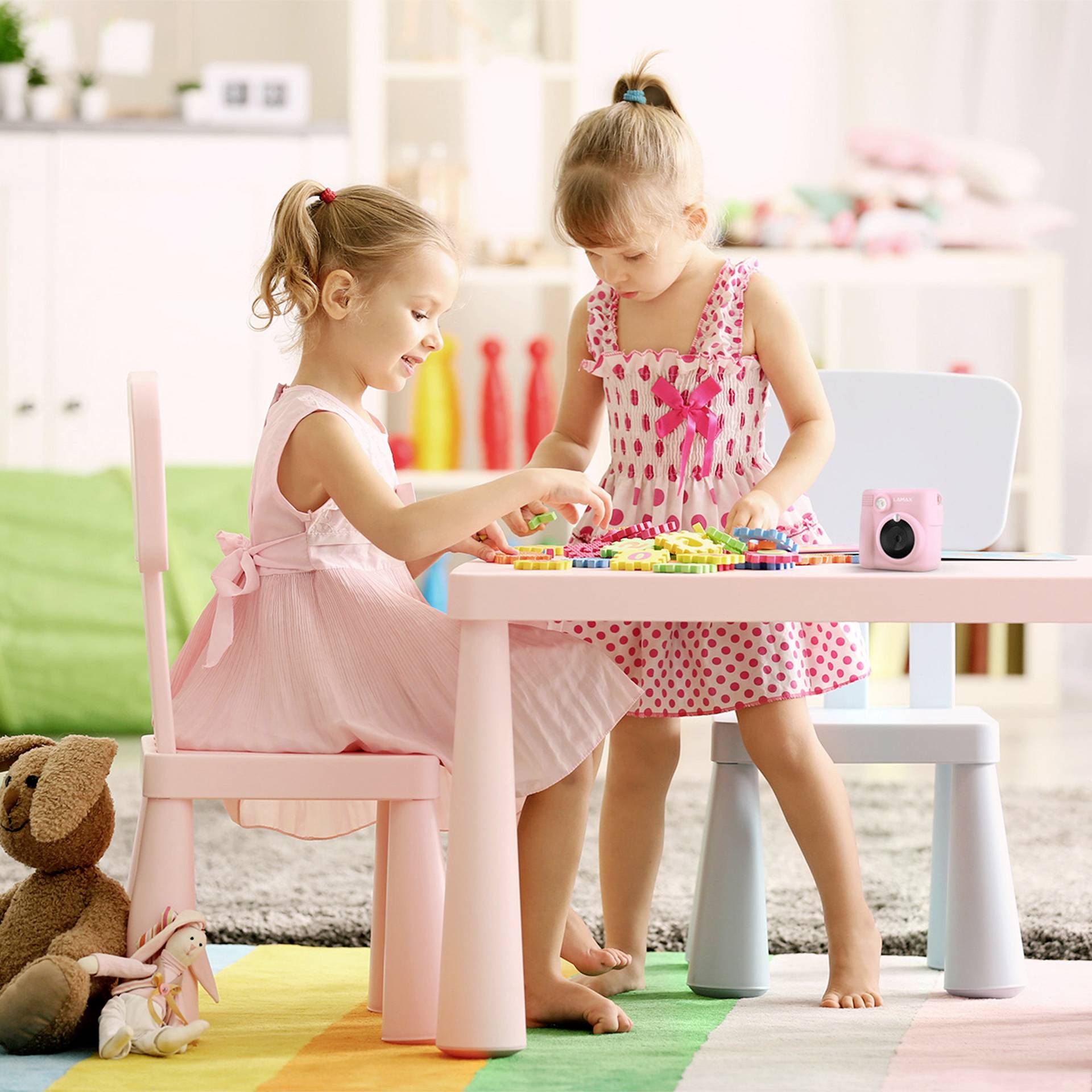 Two little girls are playing with building blocks at a colourful table in a playroom. Toys are visible in the background.