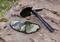 Folding shovel with serrated edge and black handle, resting on rocks. Camouflage cover beside it. Ideal for outdoor operations.