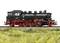 A black steam locomotive with red wheels stands on a railway track, surrounded by green grass. It is intricately and realistically designed.
