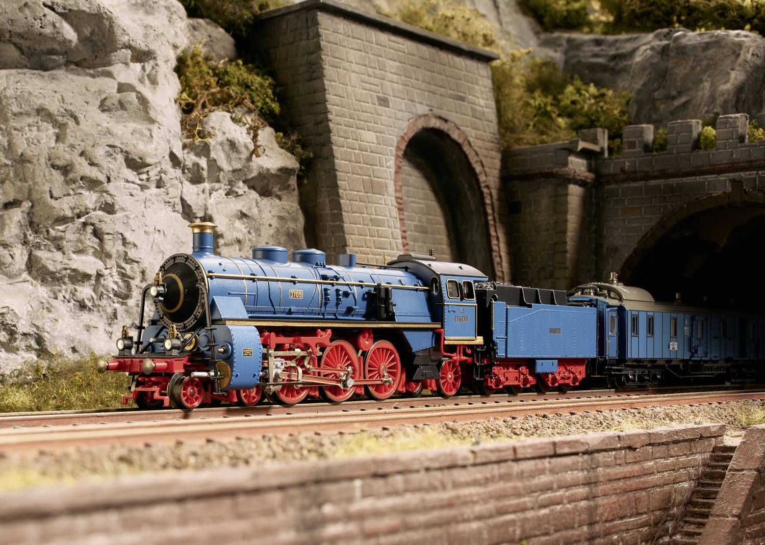 A blue and red steam locomotive emerges from a tunnel on a model railway track. Background features rocks and a brick-built tunnel.