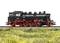 Steam locomotive on a railway track, side profile. Black boiler structure with red chassis. Surrounded by grass.