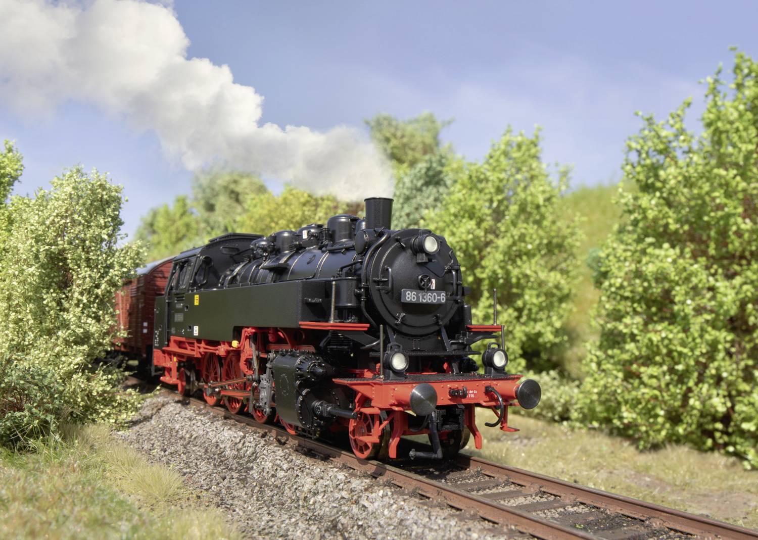 A black steam locomotive travels along rails through a green landscape, surrounded by trees and bushes, with smoke billowing from its chimney.