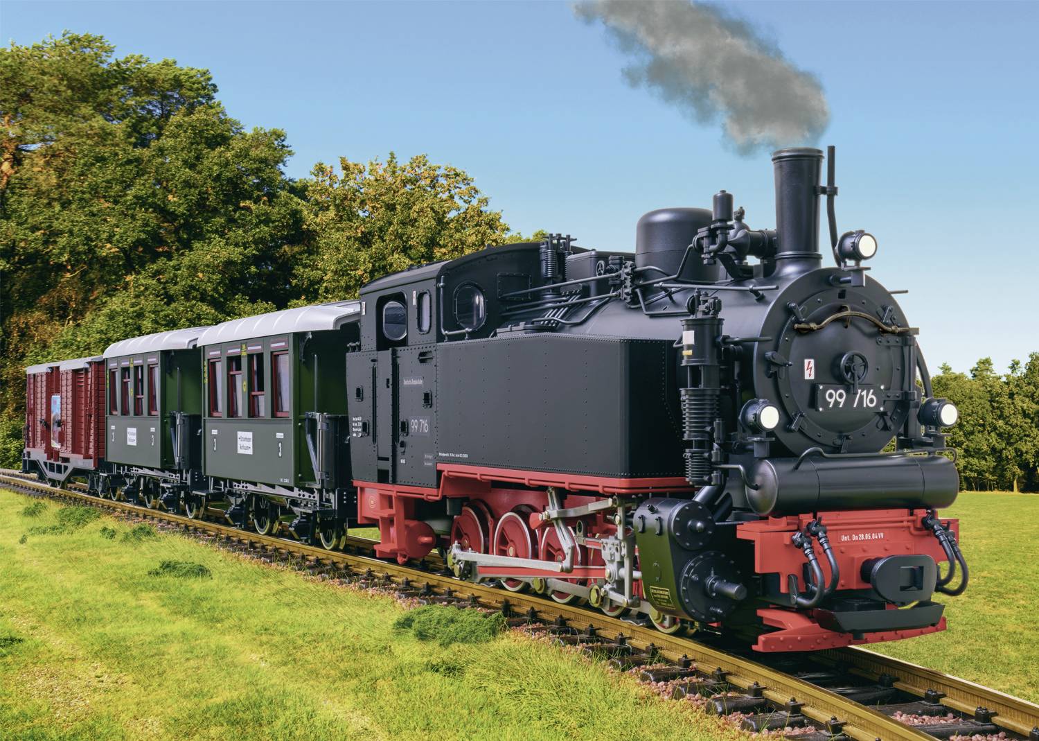 Black steam locomotive with a red undercarriage travels across a green meadow. Two historic carriages follow, with trees in the background.