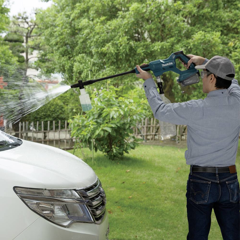 A person is cleaning the windscreen of a white car with a pressure washer outdoors, surrounded by green vegetation.