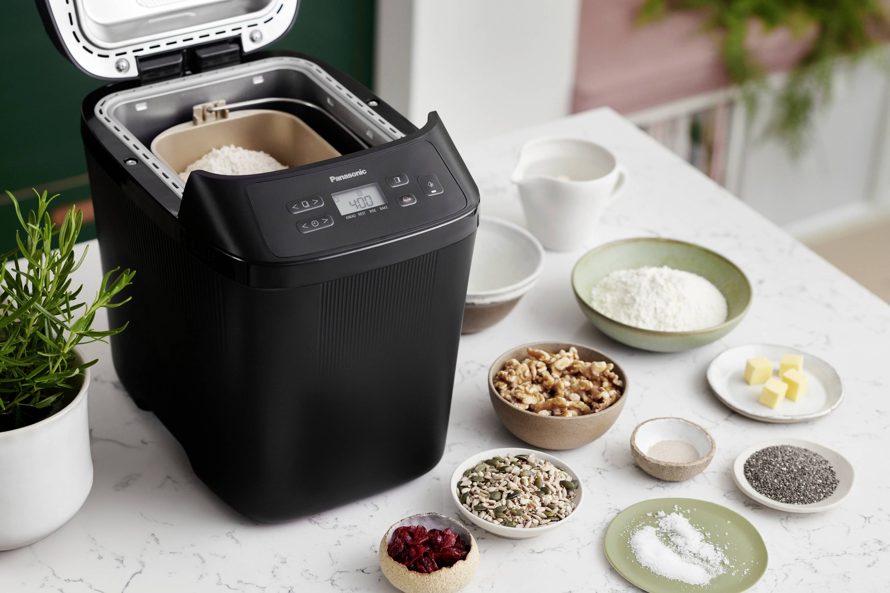 A bread maker next to ingredients like nuts, seeds, flour, butter, sugar and salt on a kitchen worktop, ready for baking bread.
