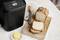 A bread machine on a kitchen table, with a chopping board beside it featuring sliced bread. A knife and butter are also visible.