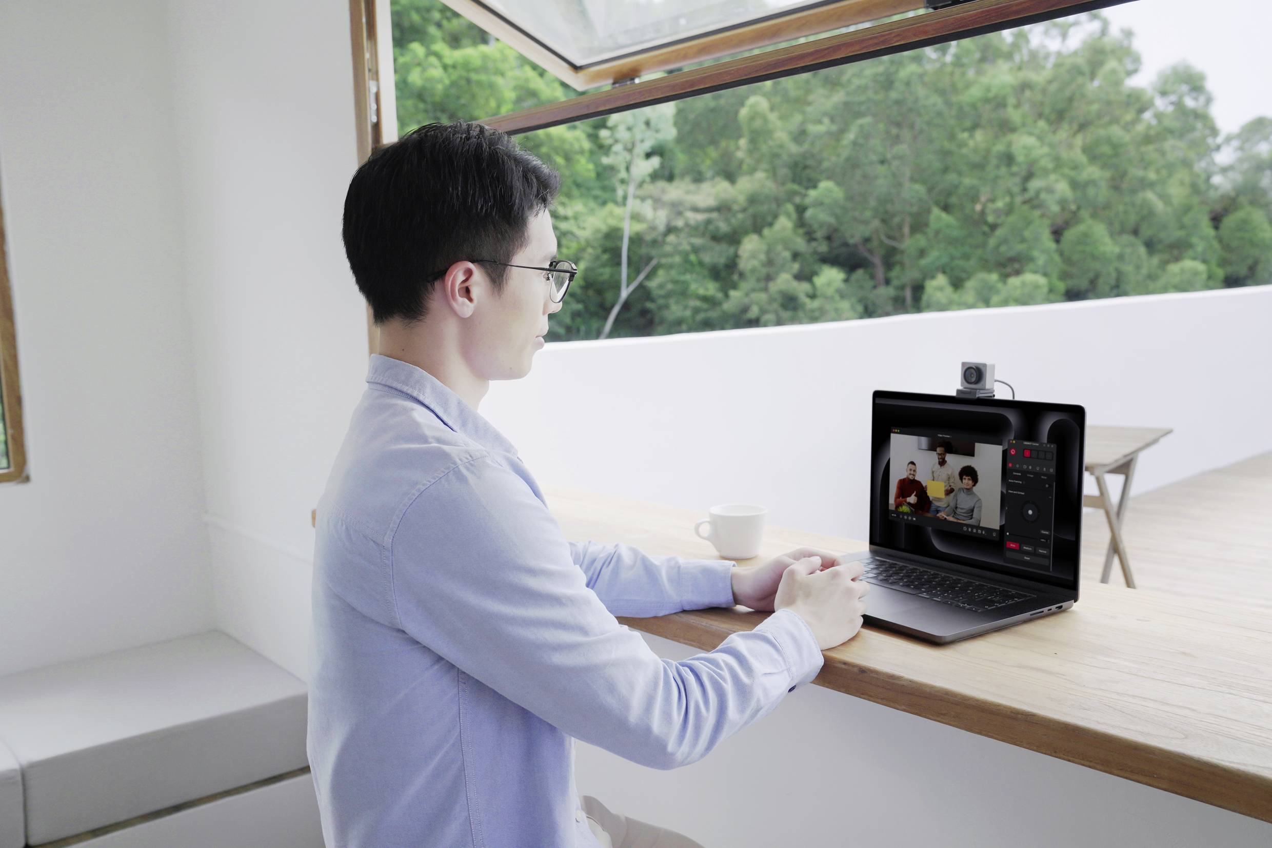 A person is sitting at a table and conducting a video call on a laptop. A green outdoor area is visible through the window.
