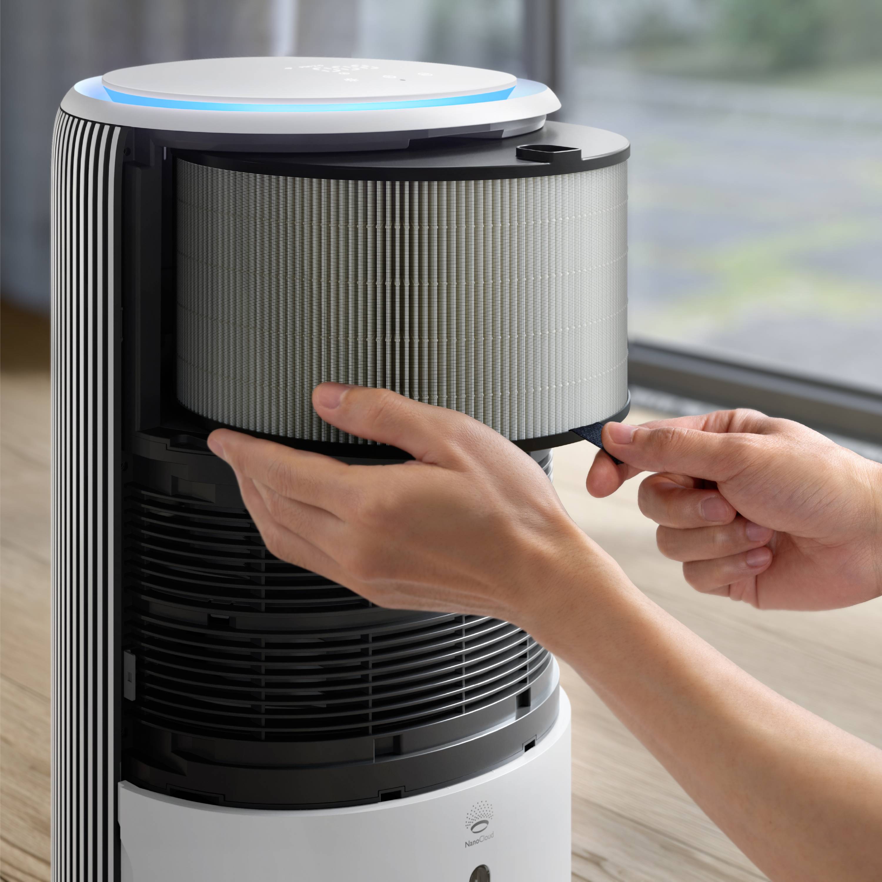 A person is replacing the filter in an air purifier that is standing next to a window. The air purifier is cylindrical and white.