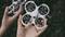 A person is holding a small, white drone with four rotor blades. The drone is hovering just above the hands. The background is out of focus.