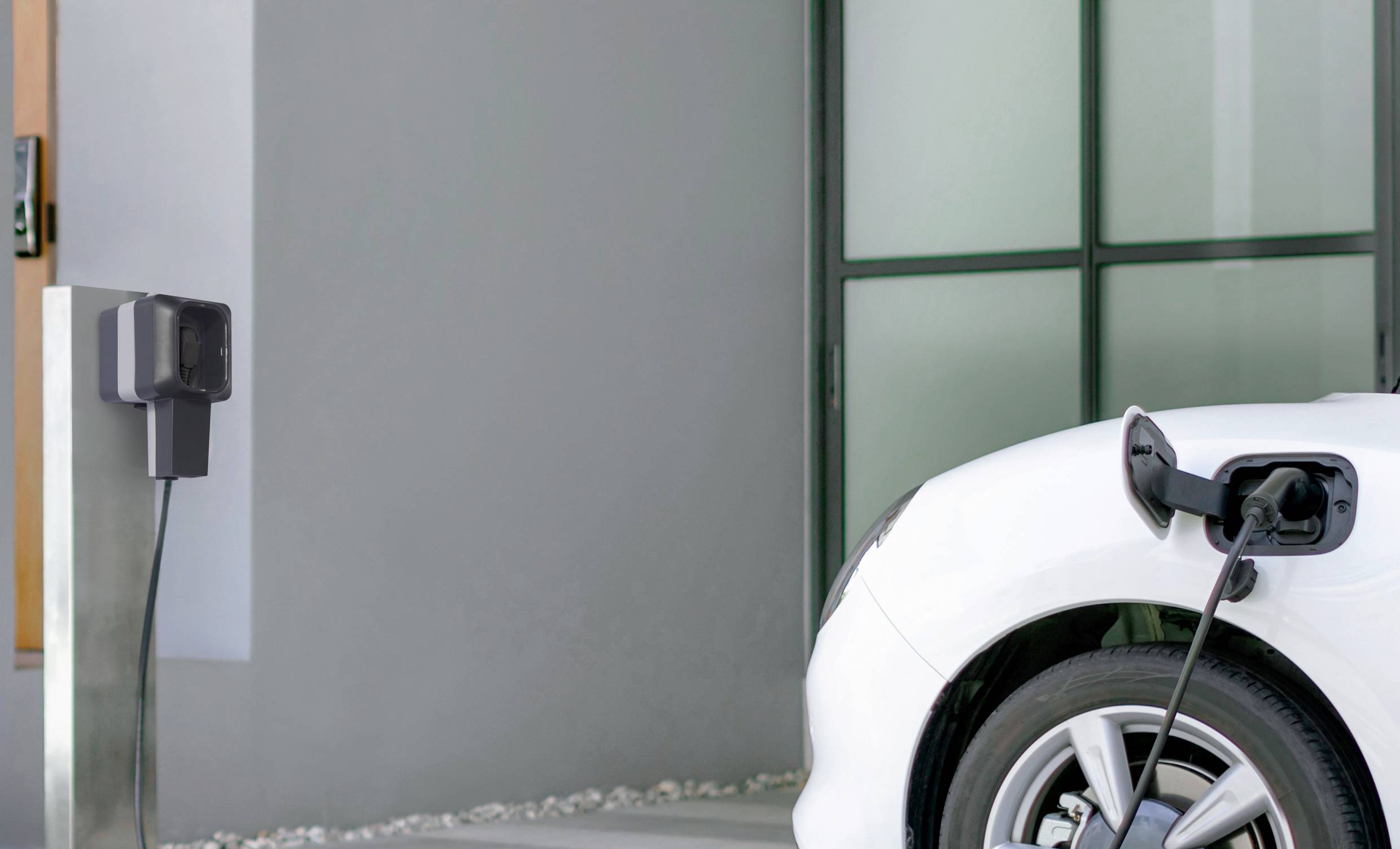 A white electric car is being charged at a charging station. The vehicle is parked in front of a modern building.