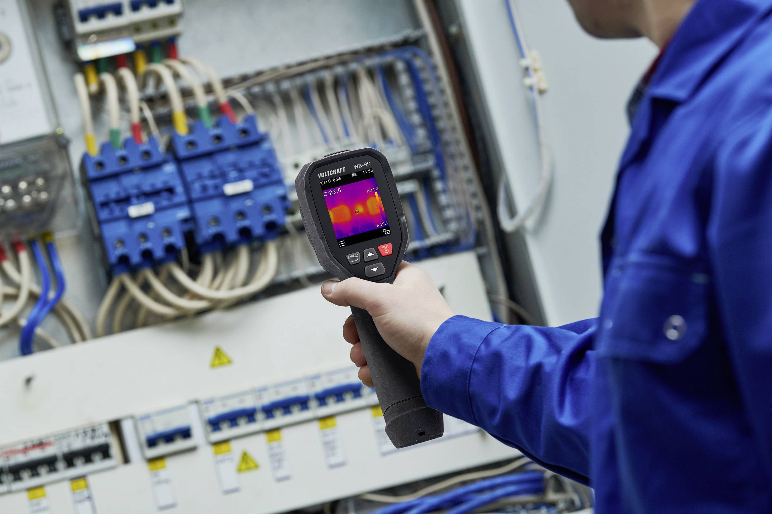 A technician in blue overalls is inspecting the cables in an electrical control cabinet using a thermal imaging camera.