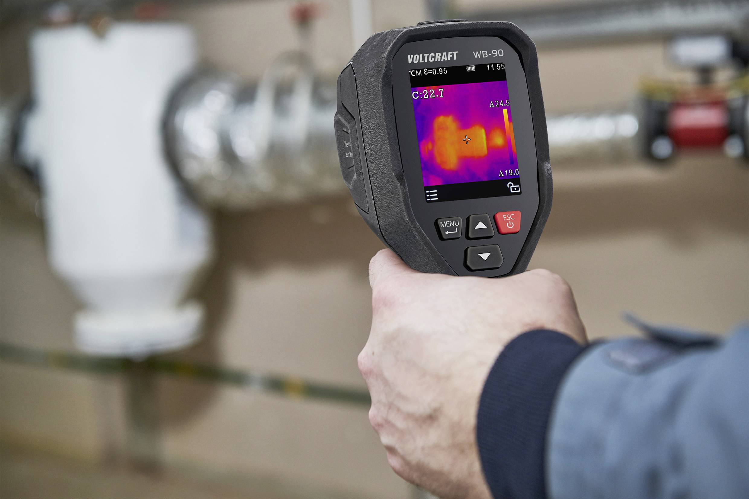 A person is holding a thermal imaging camera, measuring the temperature of a pipe. The display shows colourful temperature ranges.