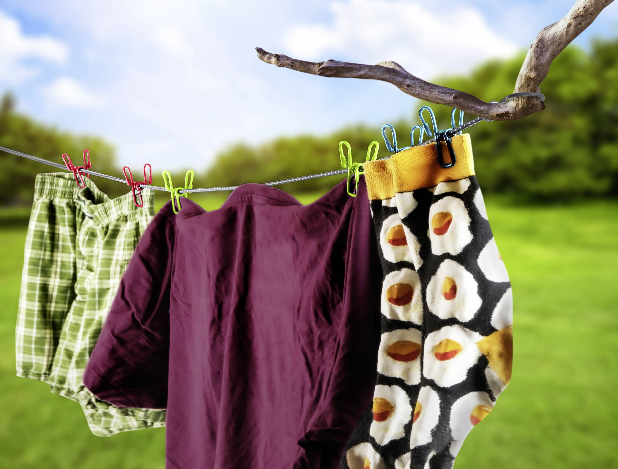 A washing line is hanging outdoors, with green checked shorts, a purple T-shirt and a multi-coloured patterned pair of trousers hanging on it.