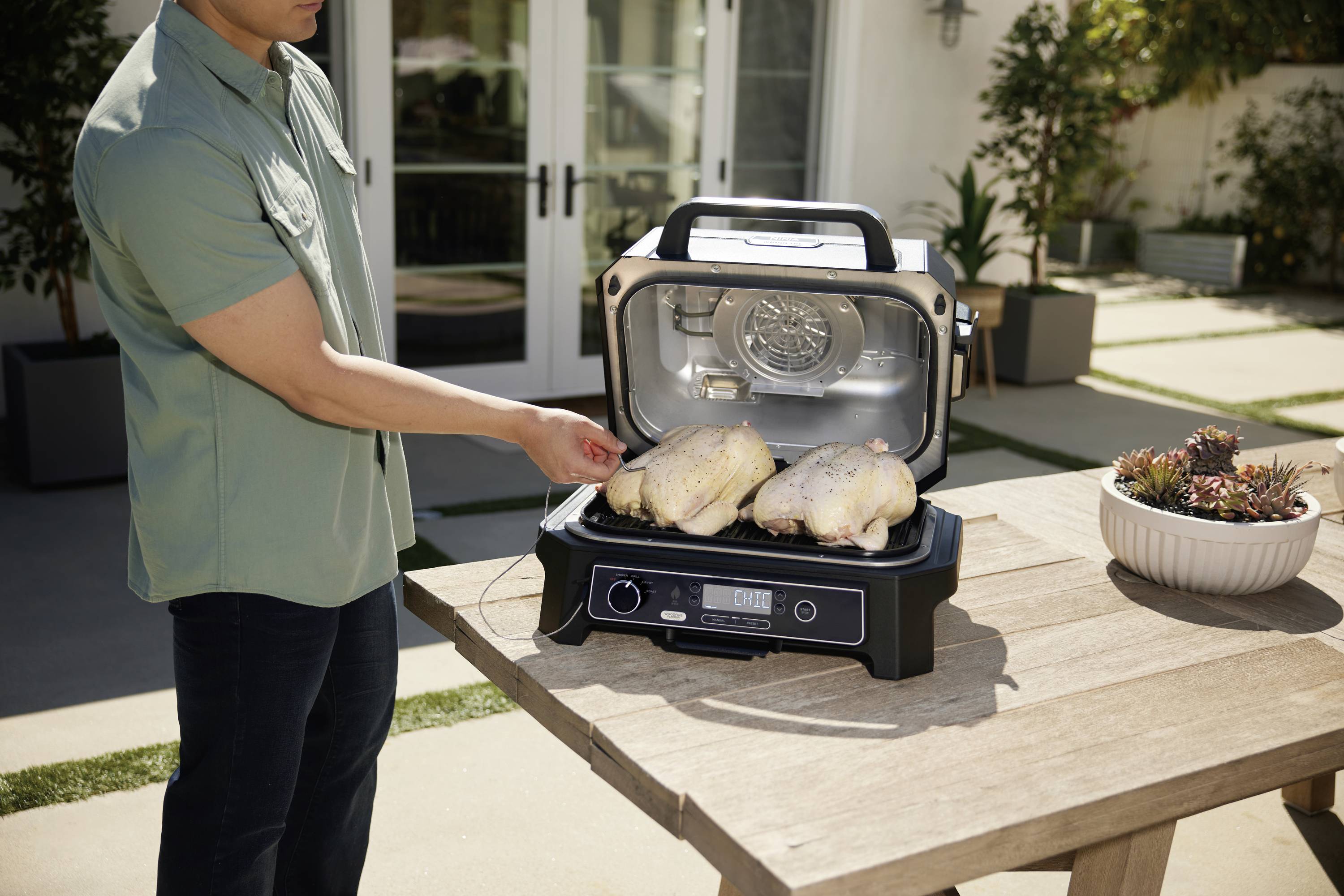 A person is grilling two chicken pieces on a modern electric barbecue outdoors. The surroundings appear sun-drenched and pleasant.