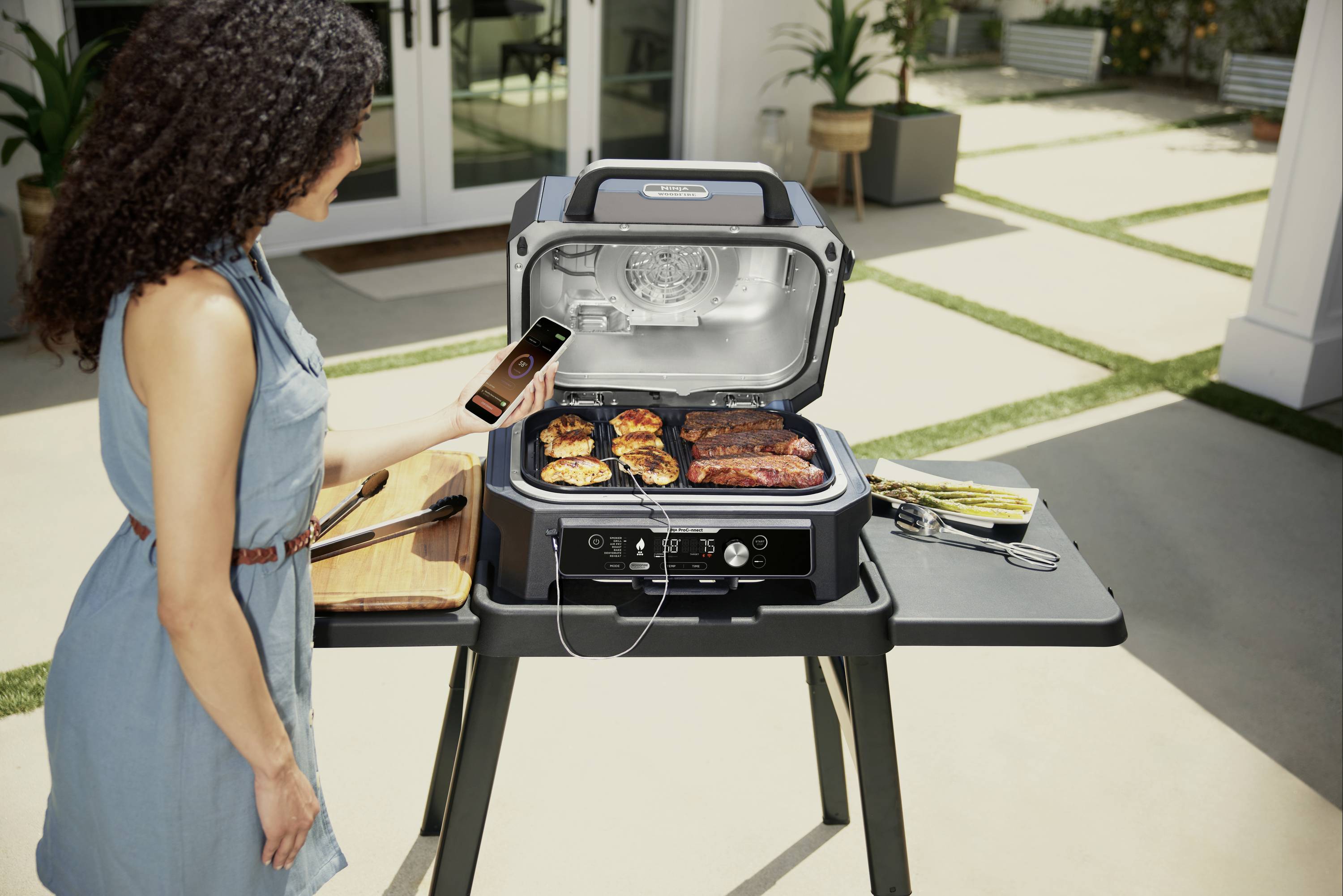 A person is grilling meat on an electric barbecue outdoors. They are holding a smartphone and monitoring the cooking process.
