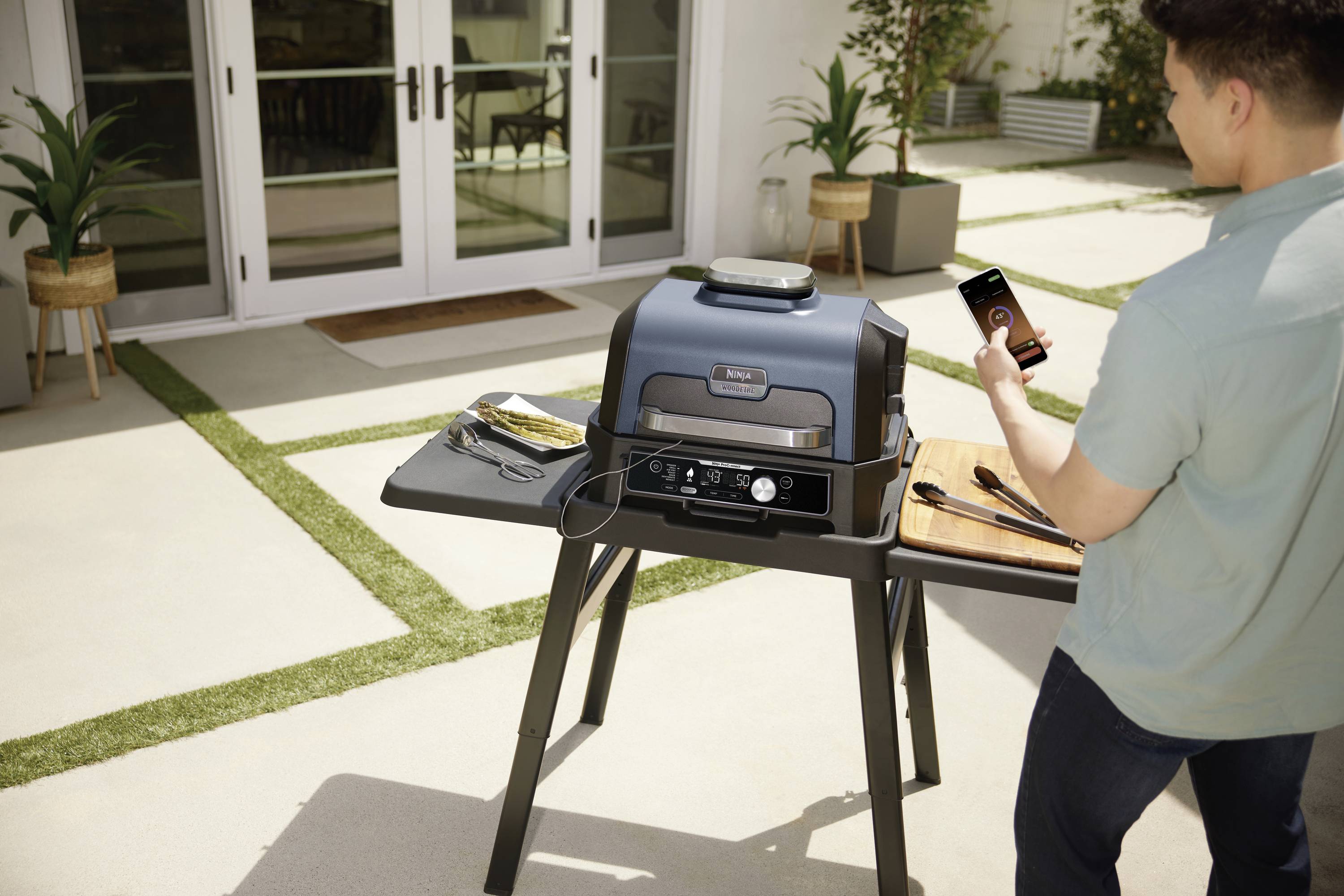 A person in a backyard is operating a modern barbecue using a mobile phone app. The grill is positioned on a table and surrounded by plants.