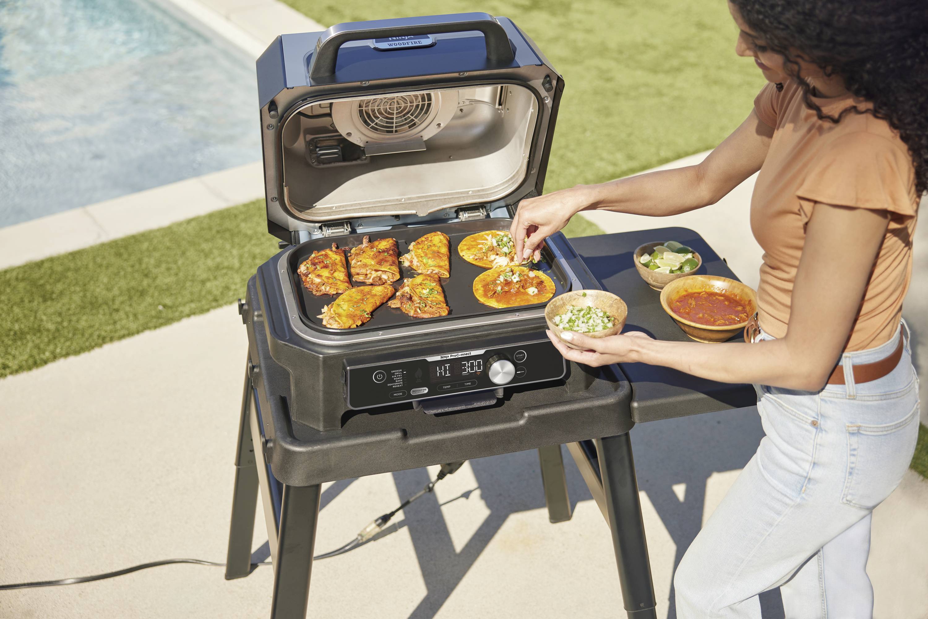 A person is grilling vegetable tacos on an electric barbecue next to a swimming pool. One hand is sprinkling herbs onto the tacos, with additional ingredients laid out nearby.