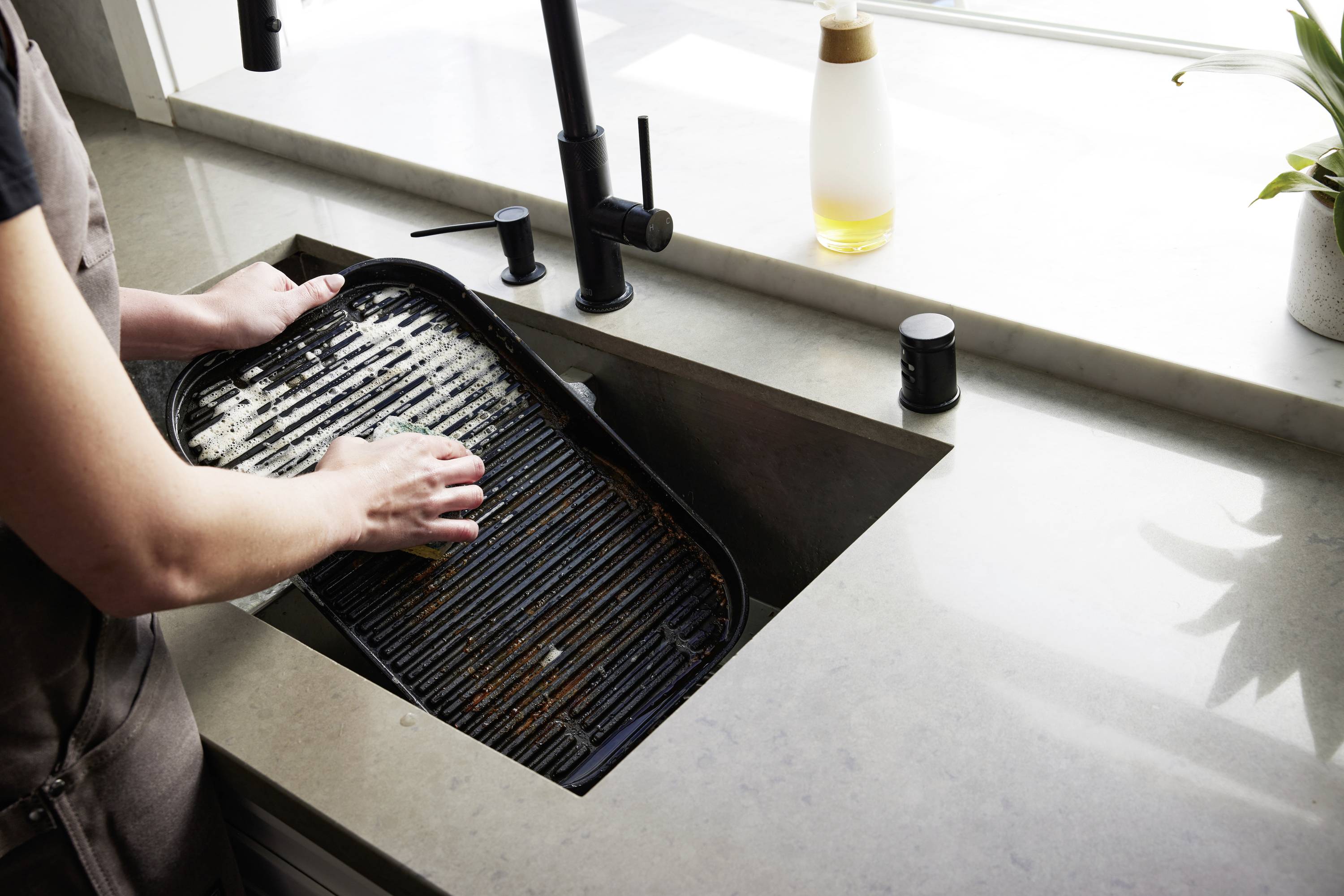 A person is washing a dirty barbecue grill in a sink. A soap bottle is sitting on the worktop.