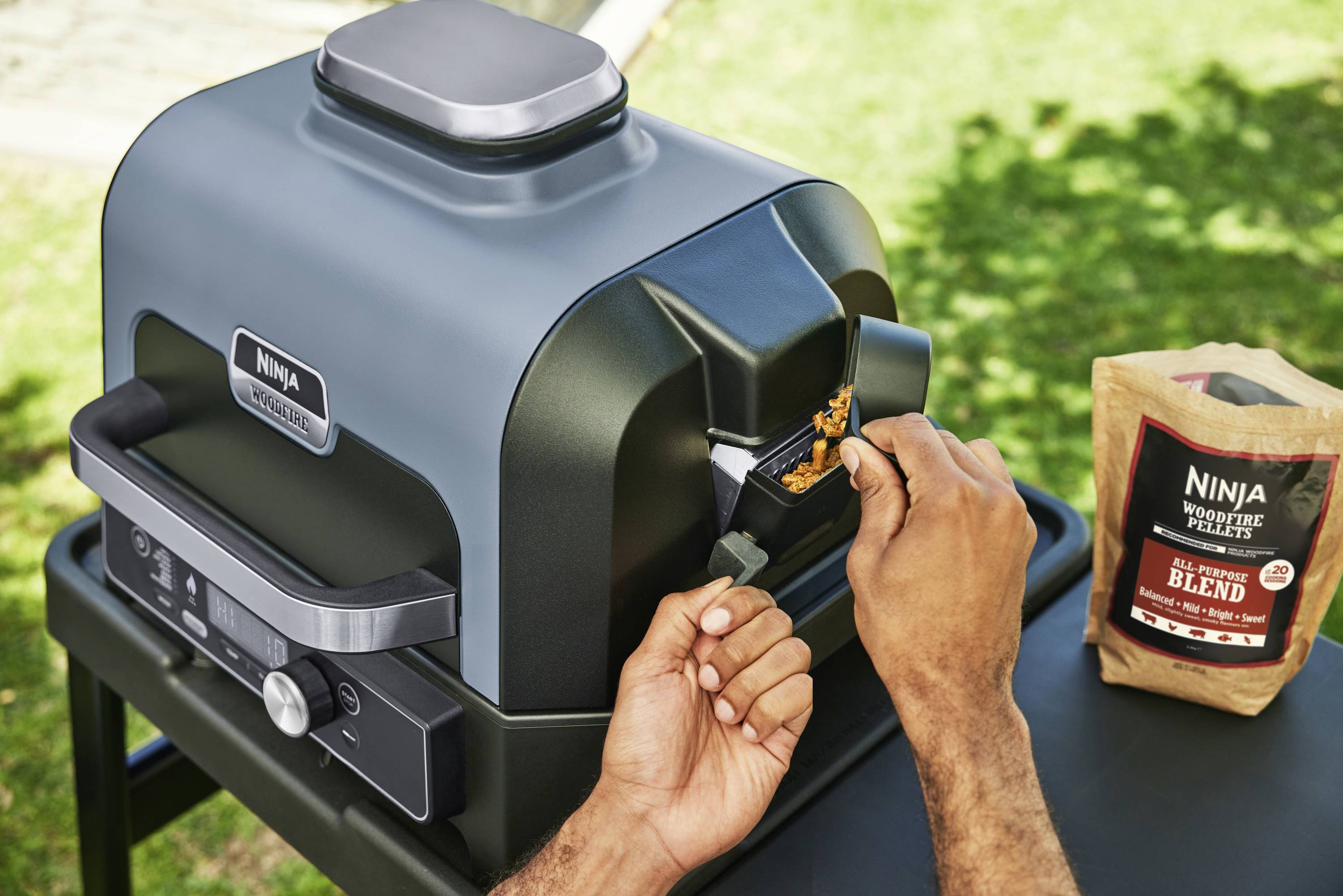 A person is filling a grill with smoking pellets in a garden setting. The grill is labelled with a 'Ninja Woodfire' tag. A packet of pellets is positioned next to the grill.