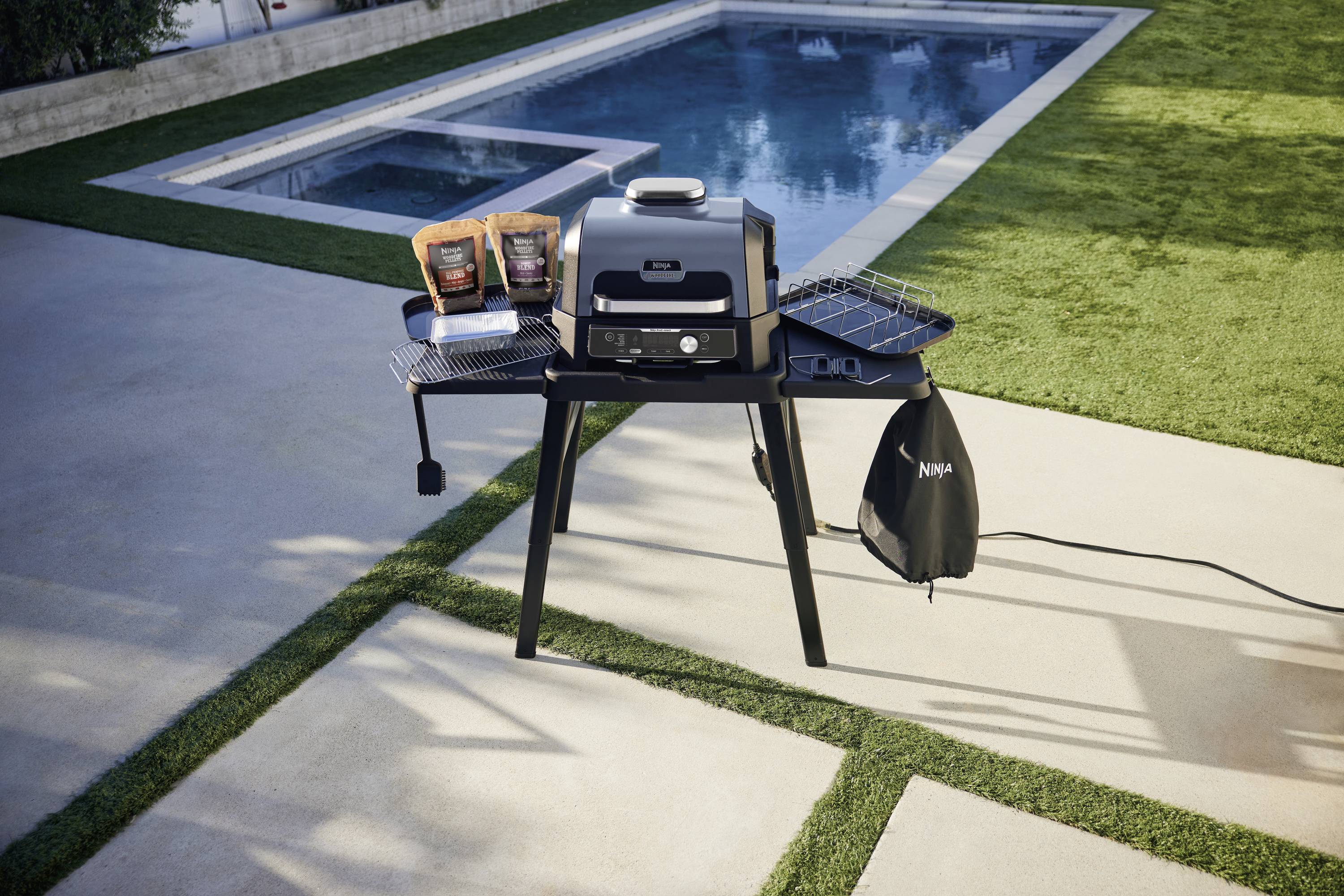 Gas barbecue in the garden next to a swimming pool, with two packets of crisps and a barbecue cover on the side surfaces. Sunny weather.
