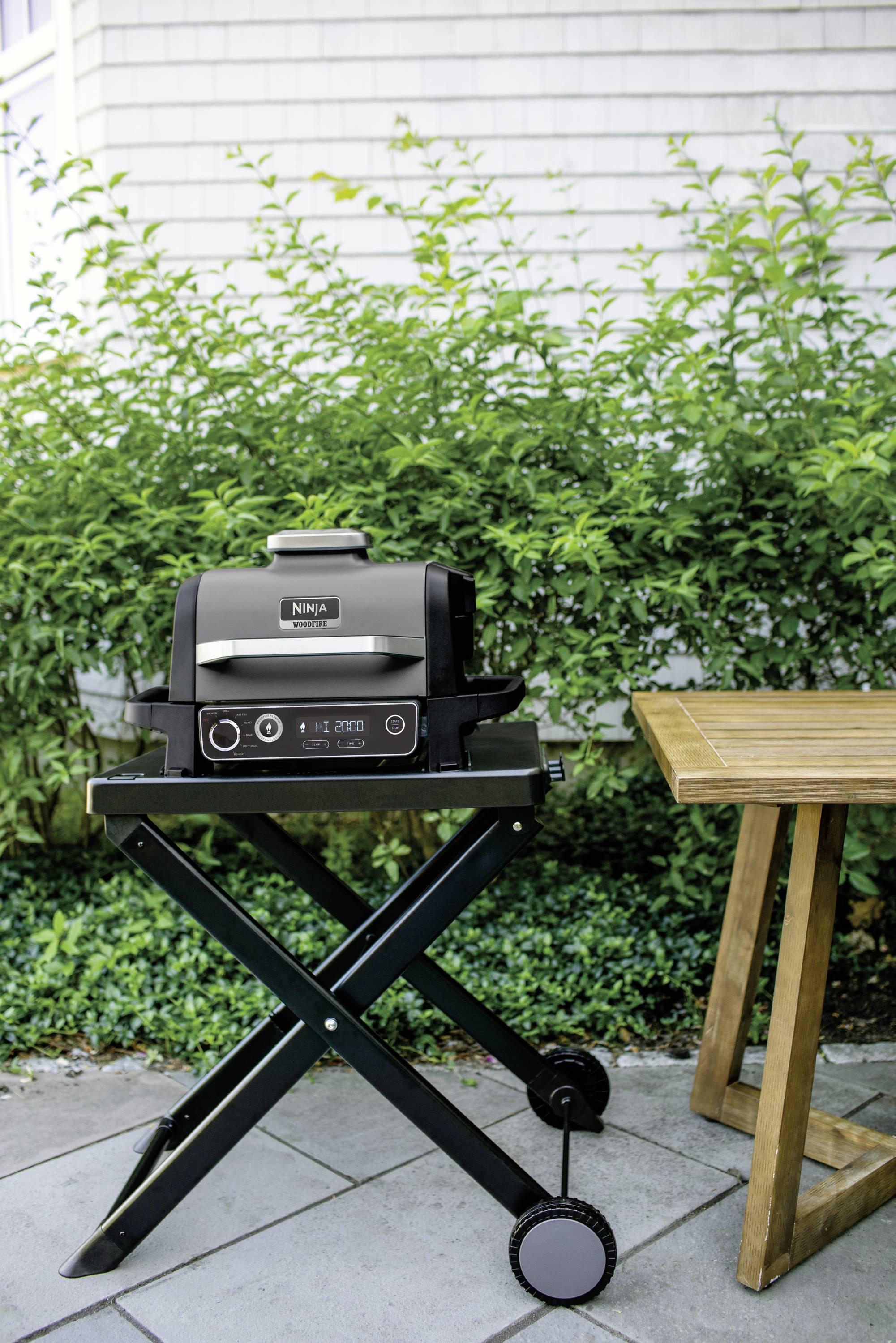 A portable barbecue stands next to a wooden table outdoors in front of a hedge. The barbecue is ready for use.