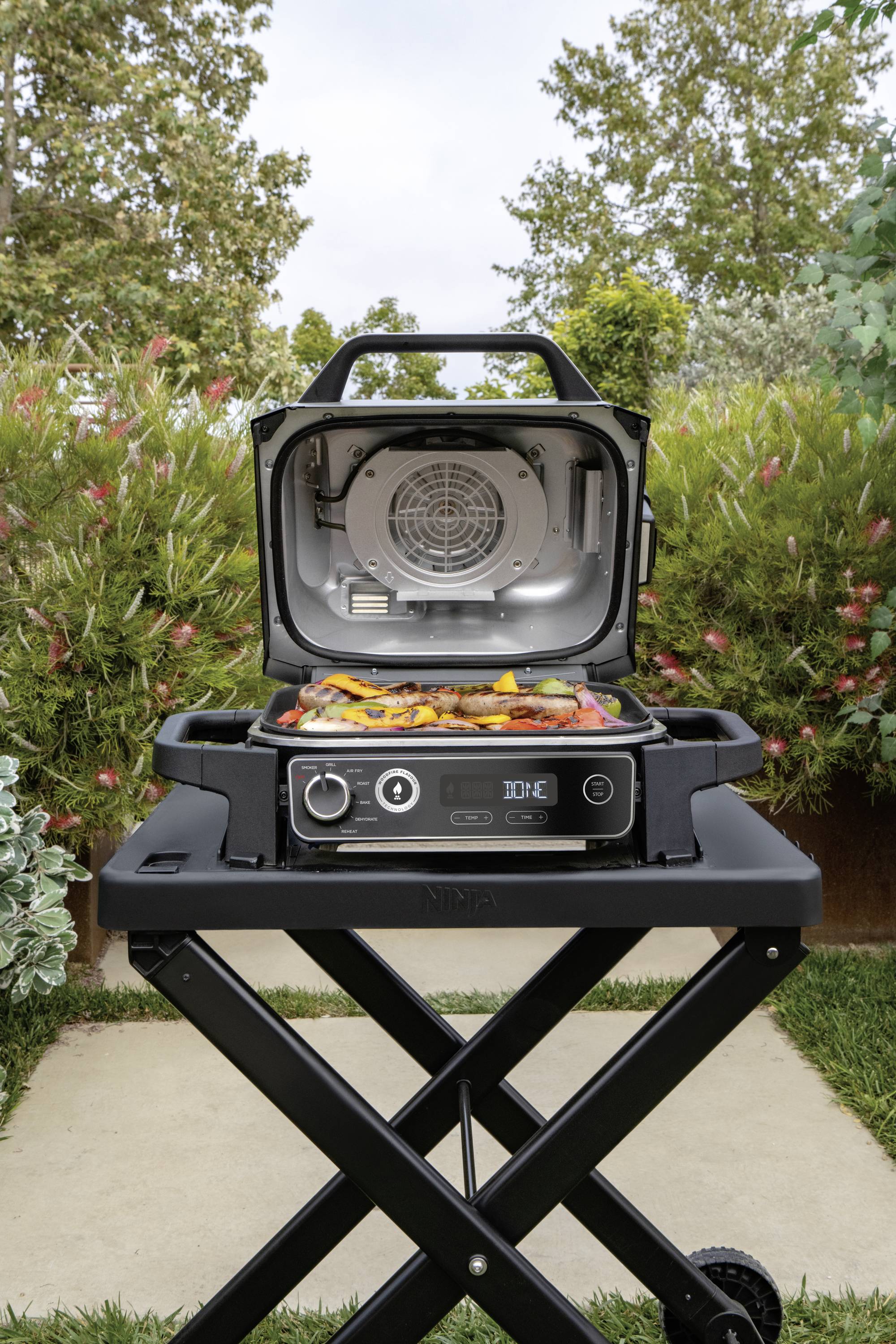 A barbecue with its lid open stands in the garden. Colourful peppers and pieces of meat are lying on the grill rack. Bushes can be seen in the background.