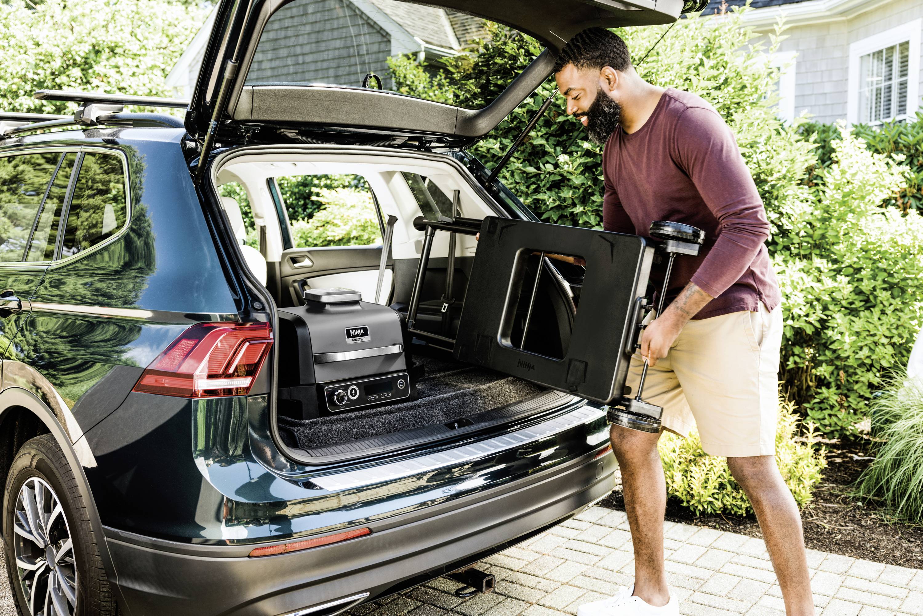 A man is loading a small barbecue into the boot of a car. The boot is open, and the car is parked in a residential area.
