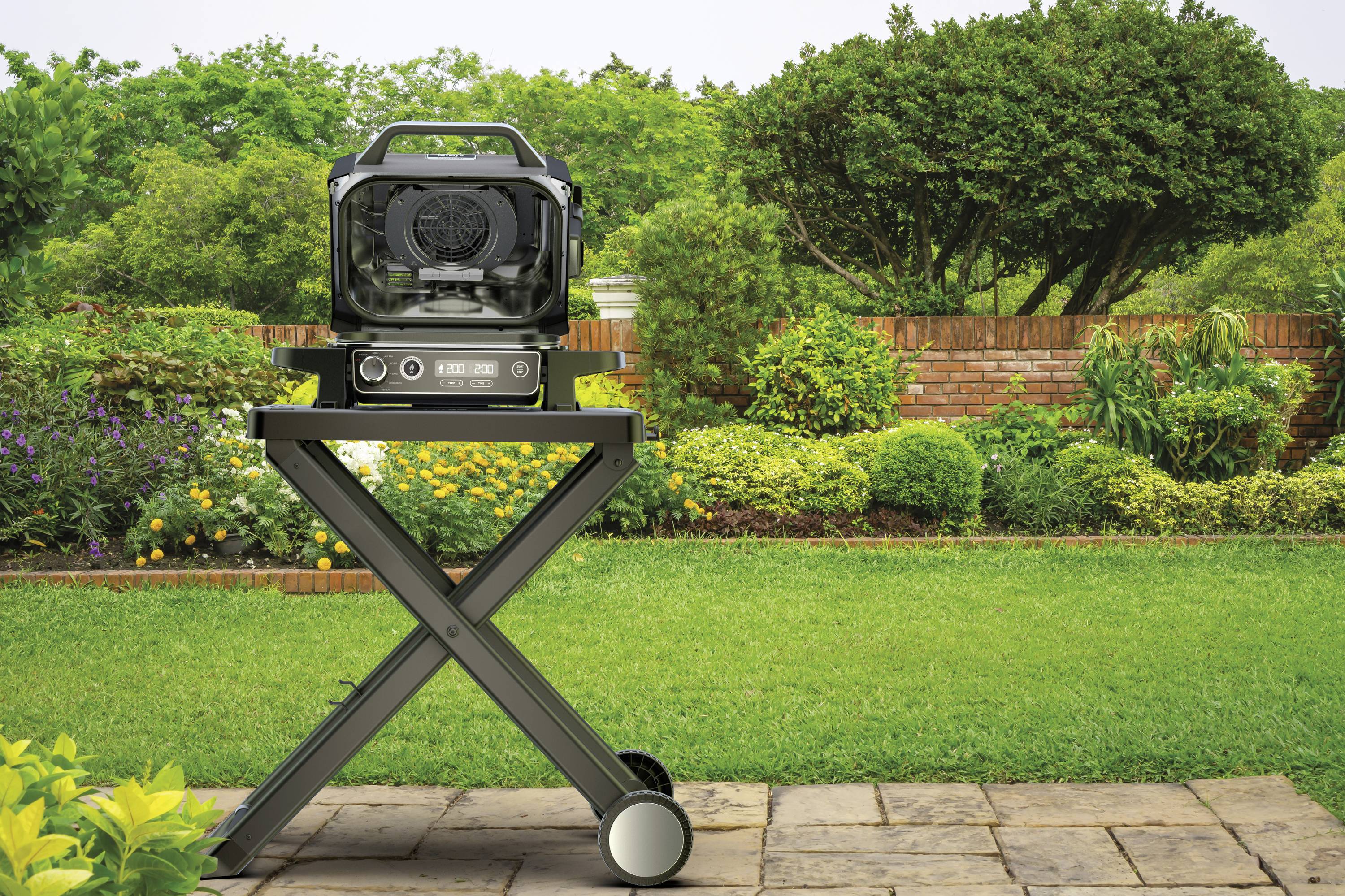 A barbecue grill sits on a trolley in the garden. In the background, green trees and colourful flowers can be seen.