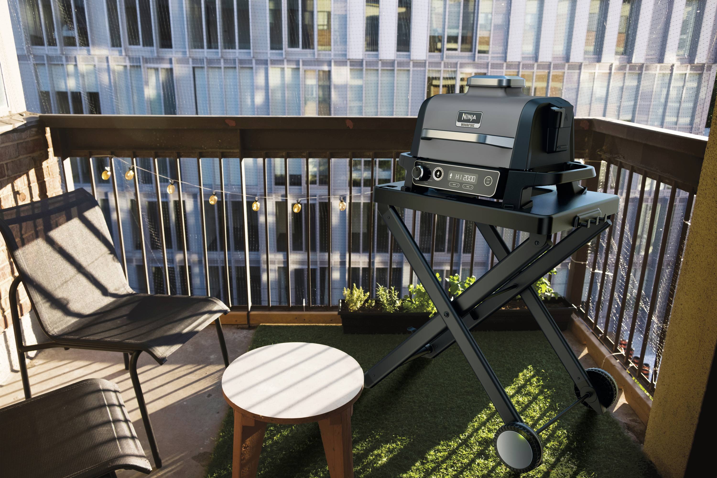 A modern barbecue stands on an urban balcony next to a small table and chair. High-rise buildings are visible in the background.