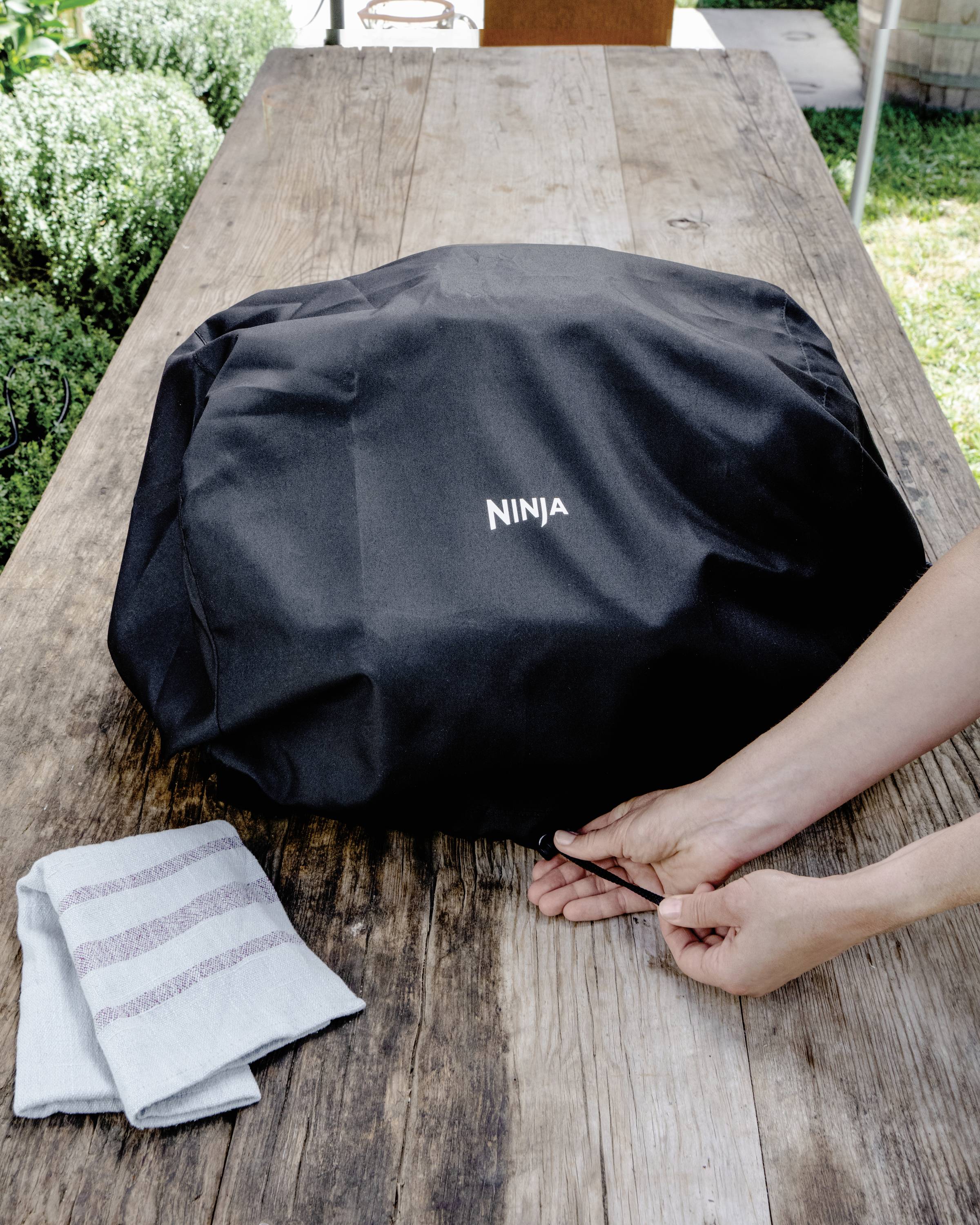 A person pulls a drawstring on a black cover with the inscription 'NINJA', placed on a wooden table next to a cloth.
