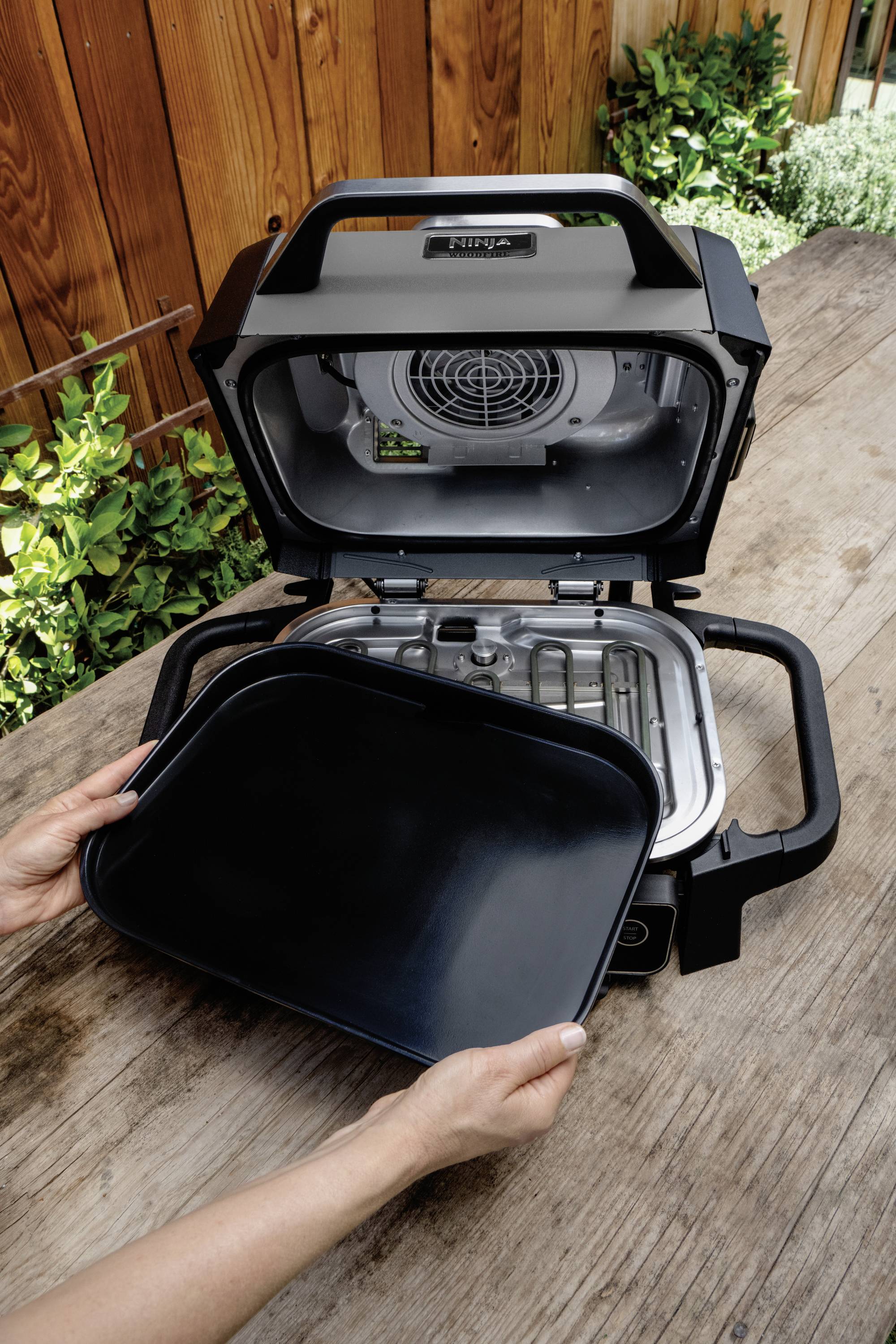 A person places a black drip tray into a portable oven on a wooden table outdoors. Plants are visible in the background.