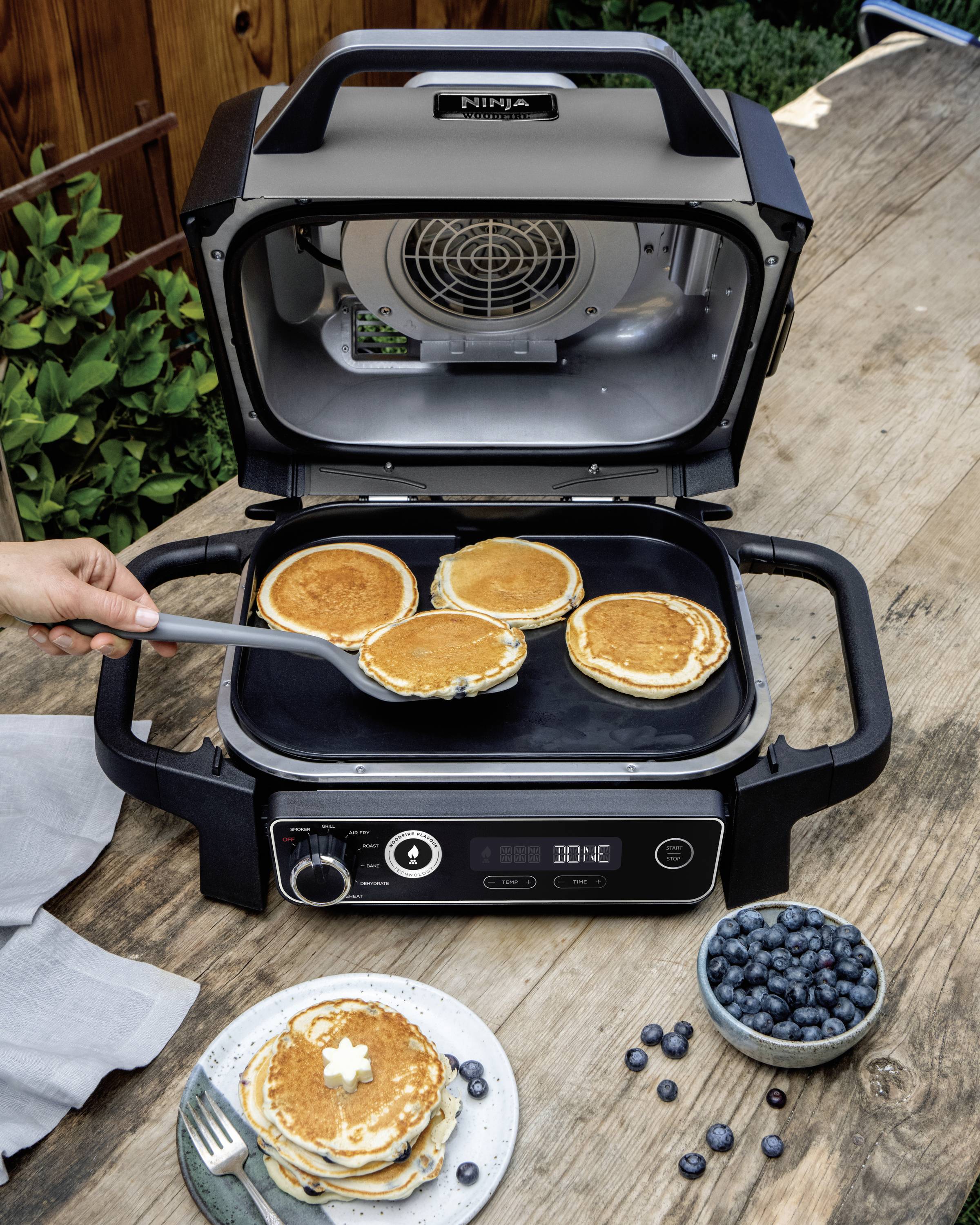 A person is lifting pancakes with a spatula from an electric griddle. On the table are a plate of pancakes and a bowl of blueberries.