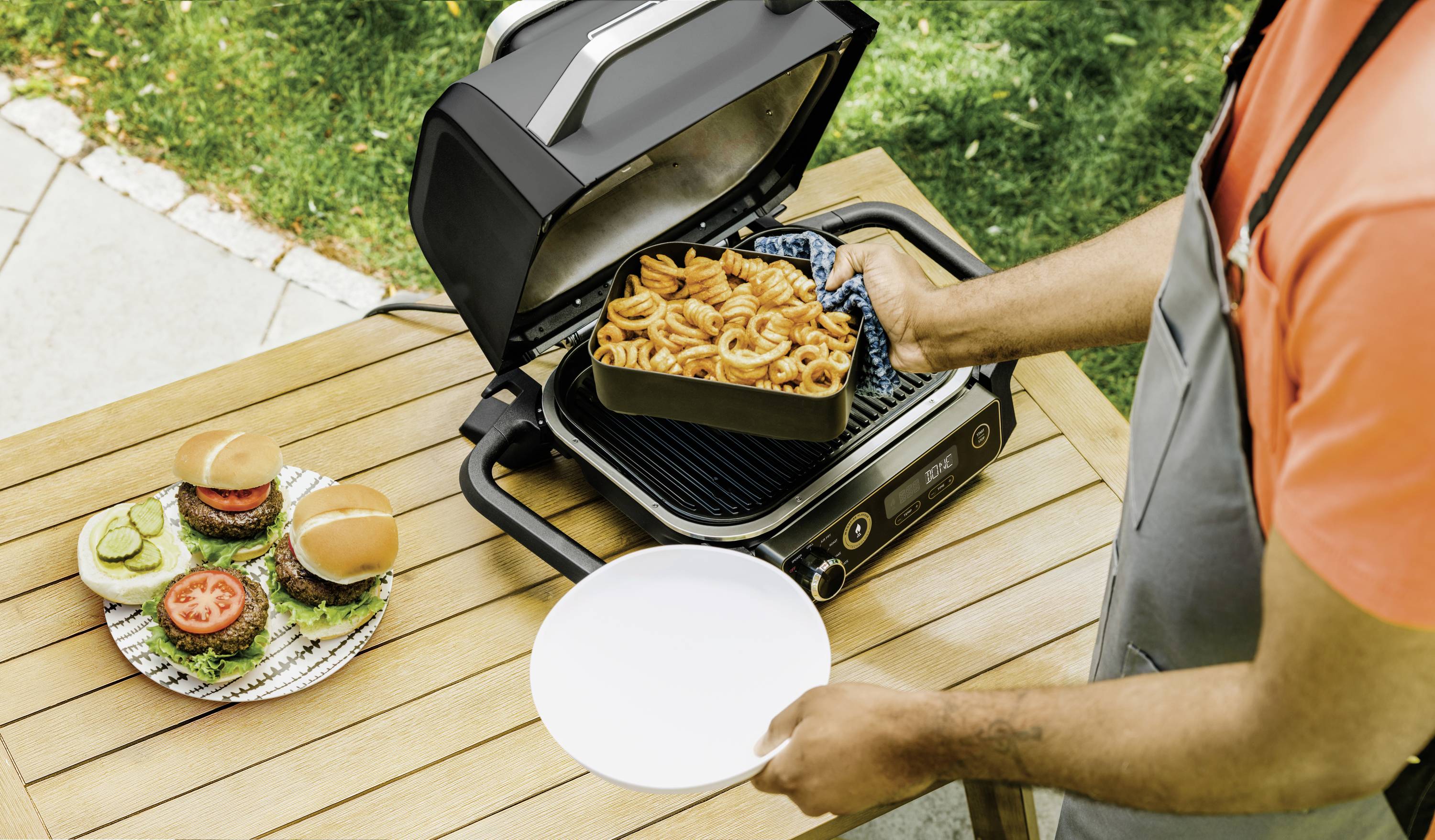 A person is grilling pasta on a tabletop grill in the garden. On the table are two burgers with lettuce, tomato, and cucumber.