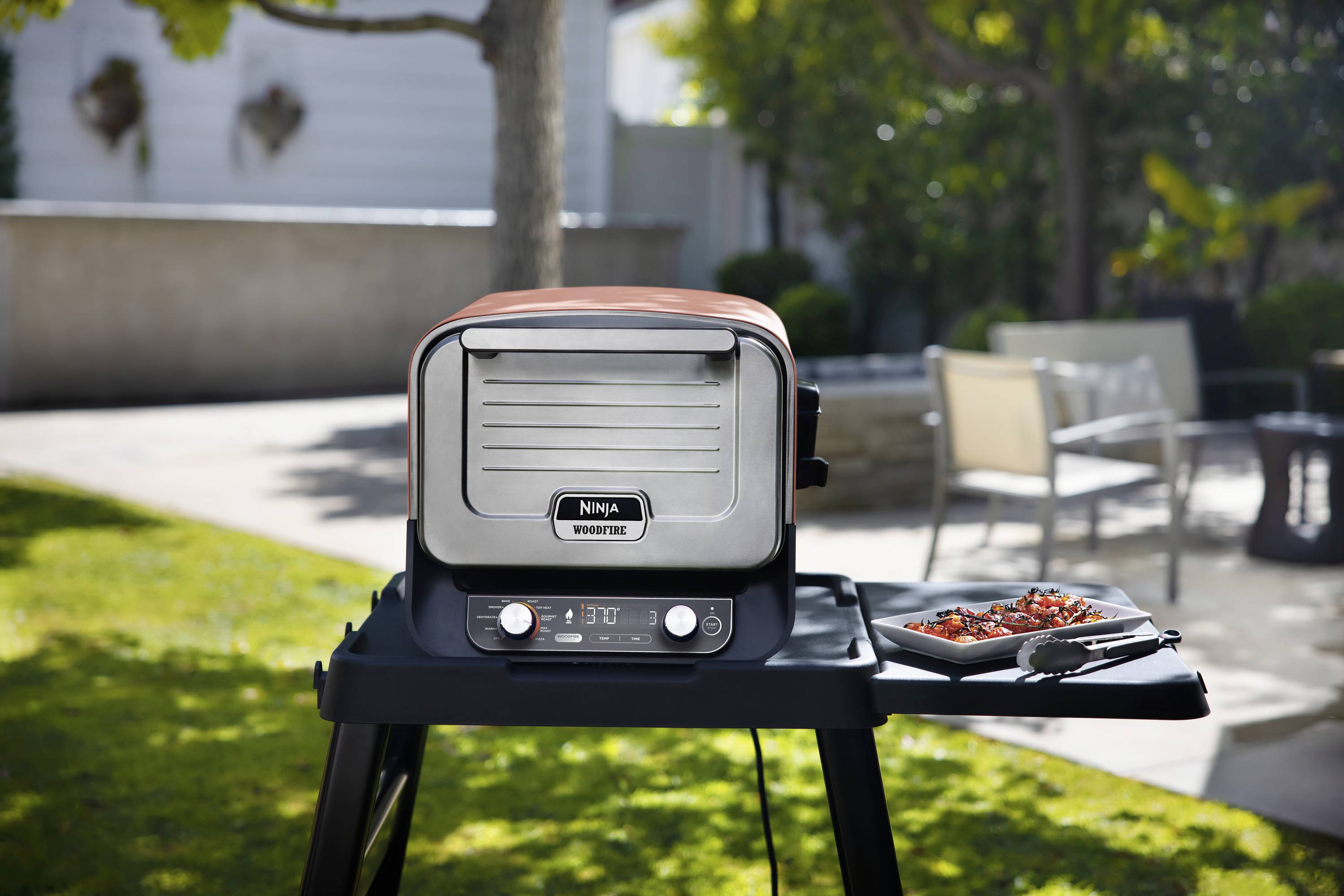 Electric barbecue in the garden, standing on a table. Next to it is a tray with marinated meat. Garden furniture is visible in the background.