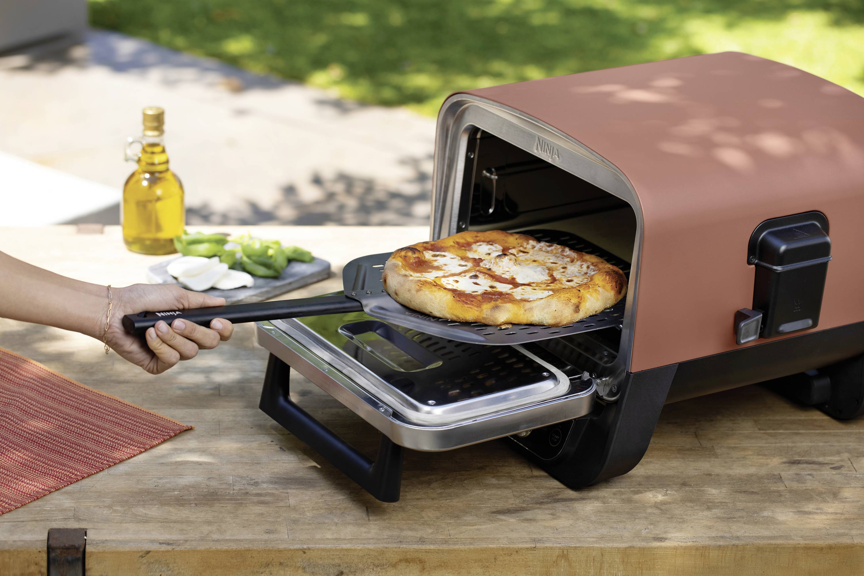 A pizza is being removed from an outdoor pizza oven with a pizza peel onto a table. A bottle of oil is lying in the background.