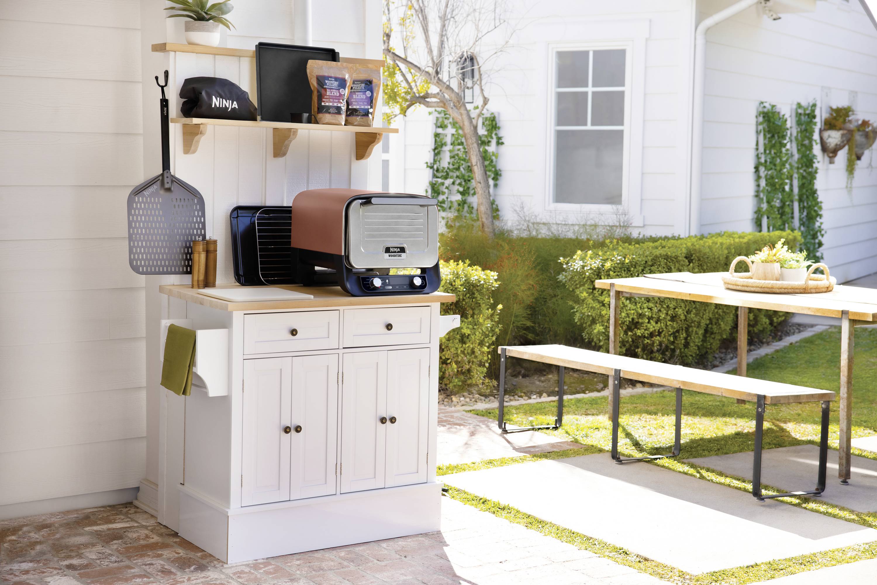Outdoor area with a white cabinet, featuring kitchen appliances and food items. In the background, a wooden picnic table on a terrace.