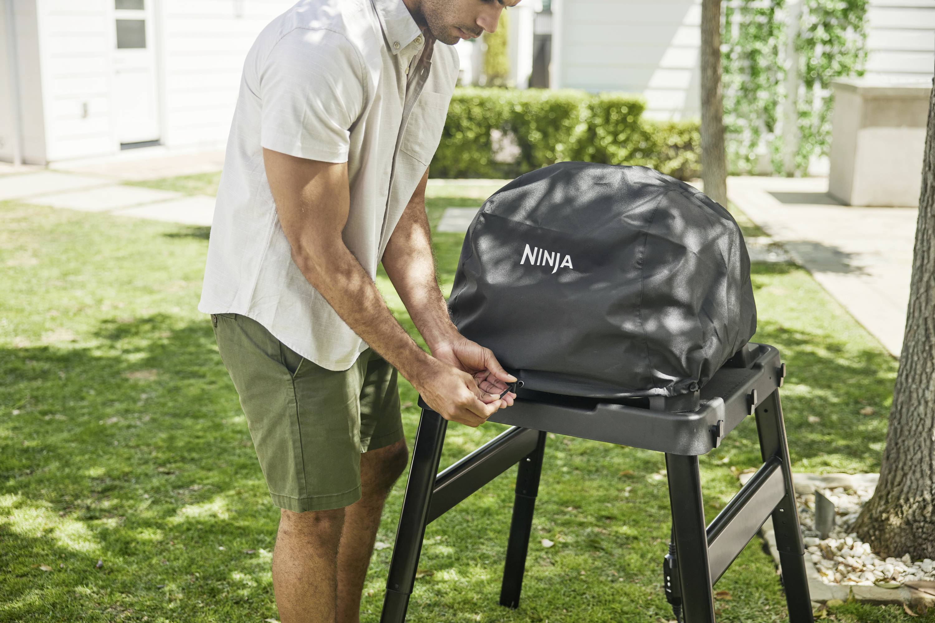 A person places a protective cover over a barbecue in the garden. The barbecue is mounted on a black stand and is set up outdoors.