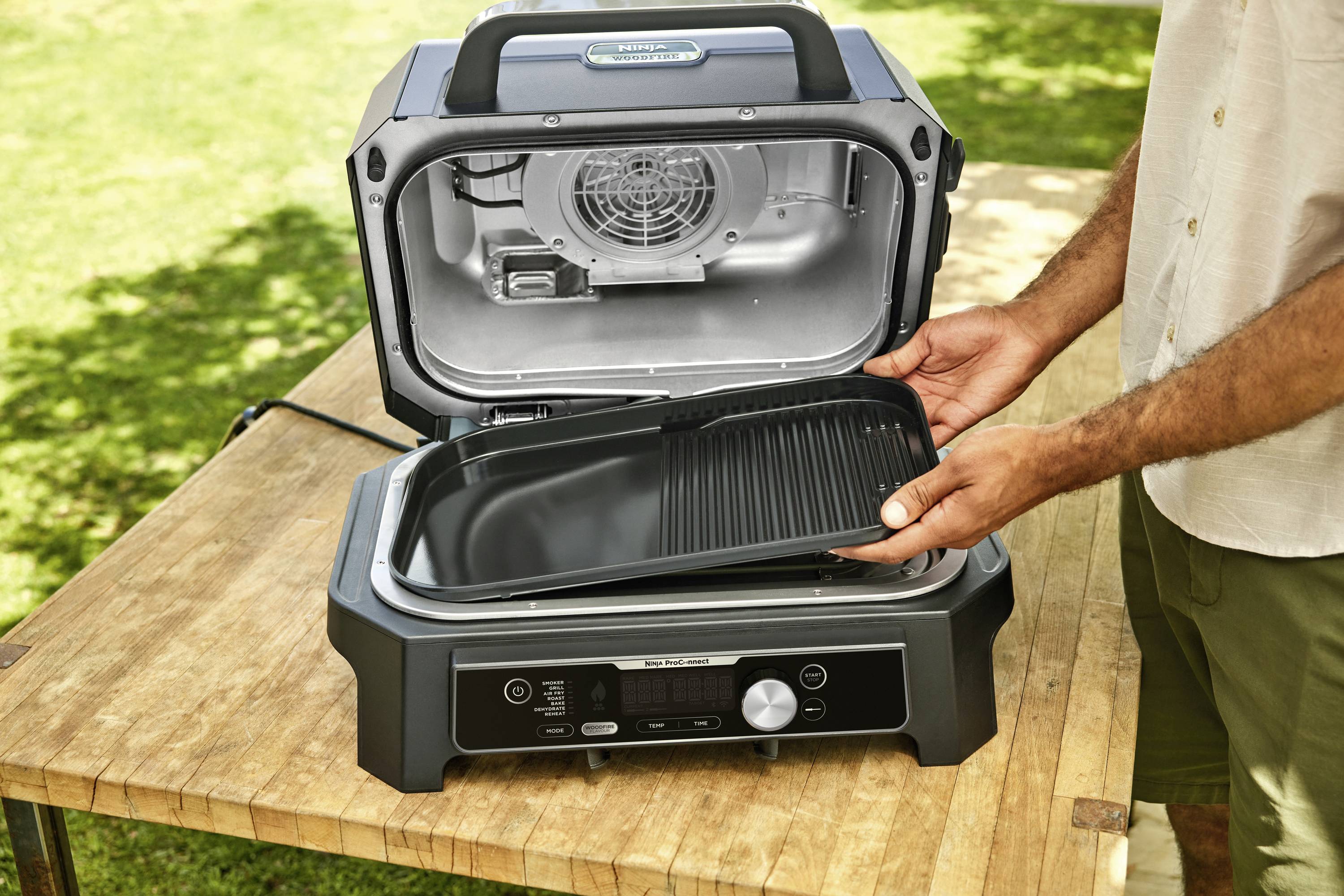 A person opens an electric barbecue with the grill rack removed on a wooden table outdoors.