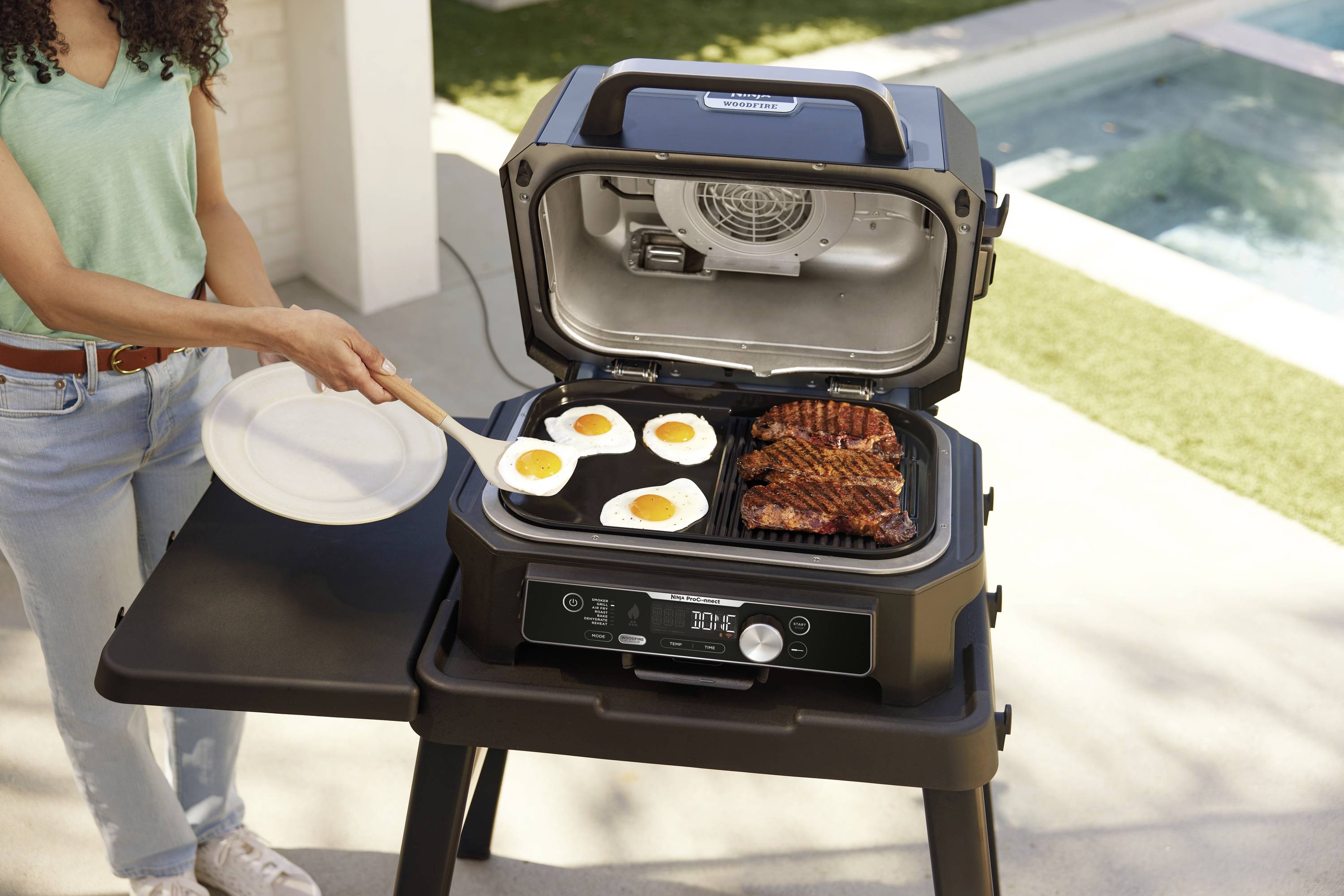 A person is standing next to an open barbecue on a patio. Four fried eggs and grilled meat can be seen on the barbecue.