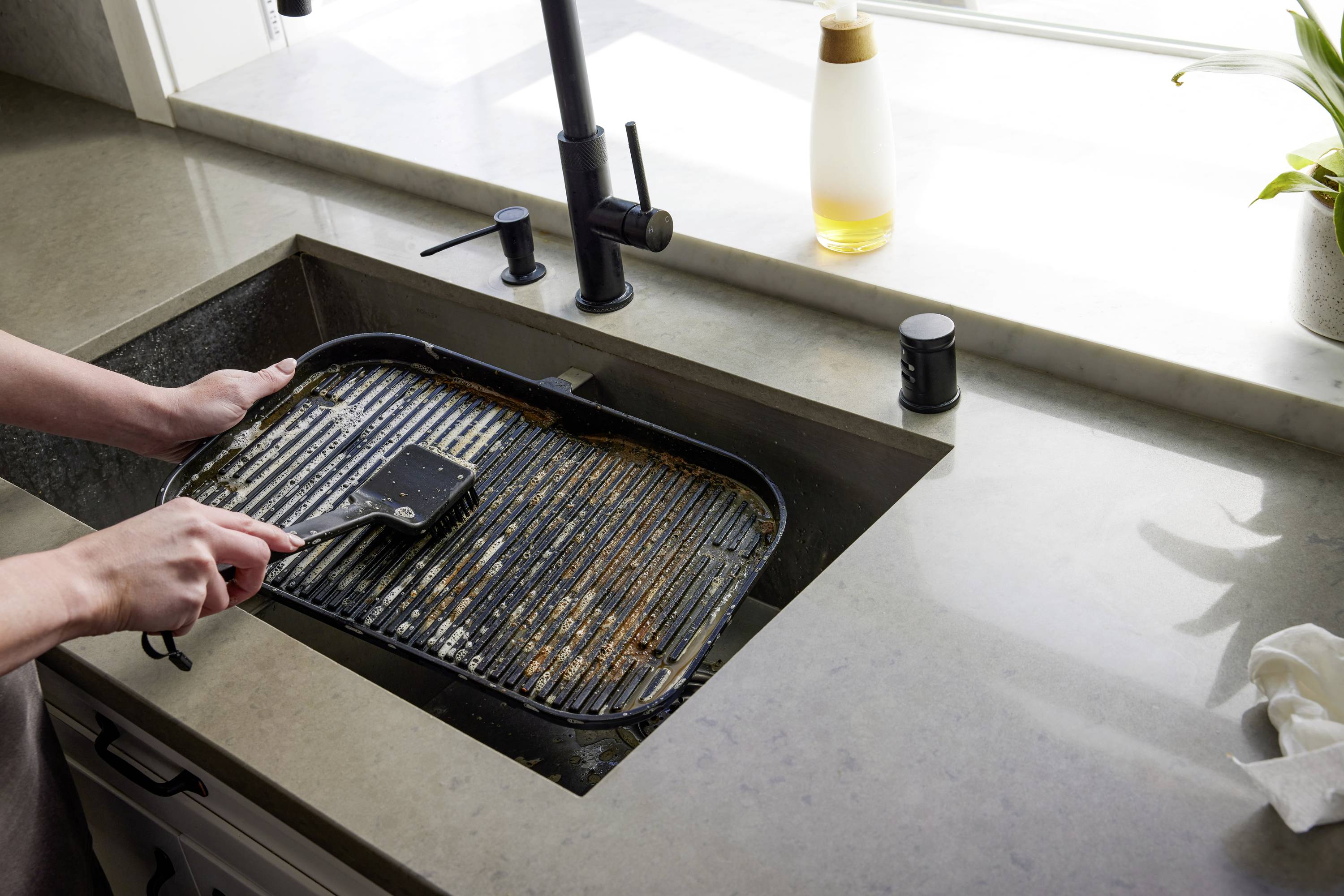 A person is cleaning a dirty baking tray in a modern kitchen sink using a scraper.