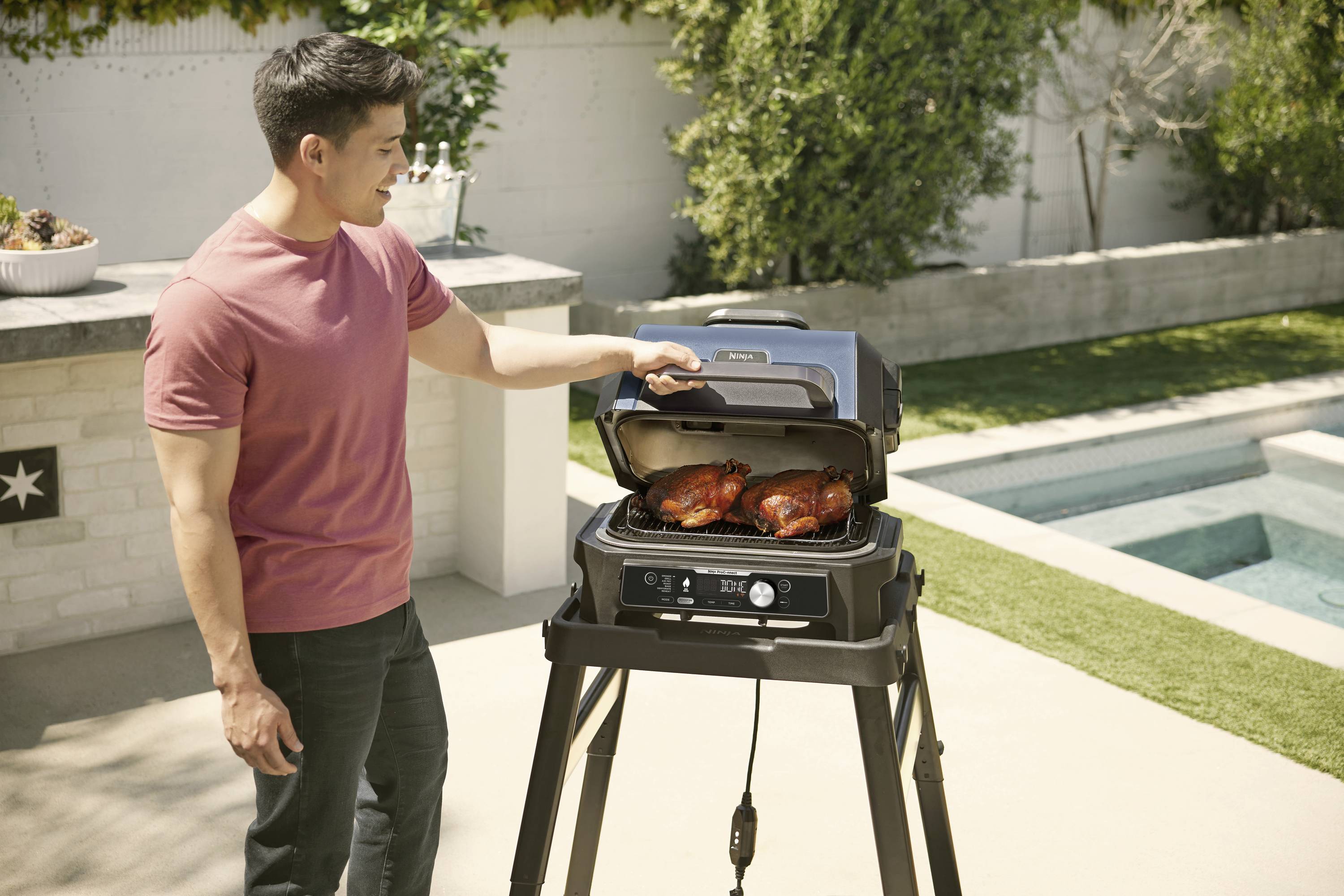A man in a garden is barbecuing chicken on a barbecue next to a swimming pool. It is sunny and he is wearing a pink T-shirt.