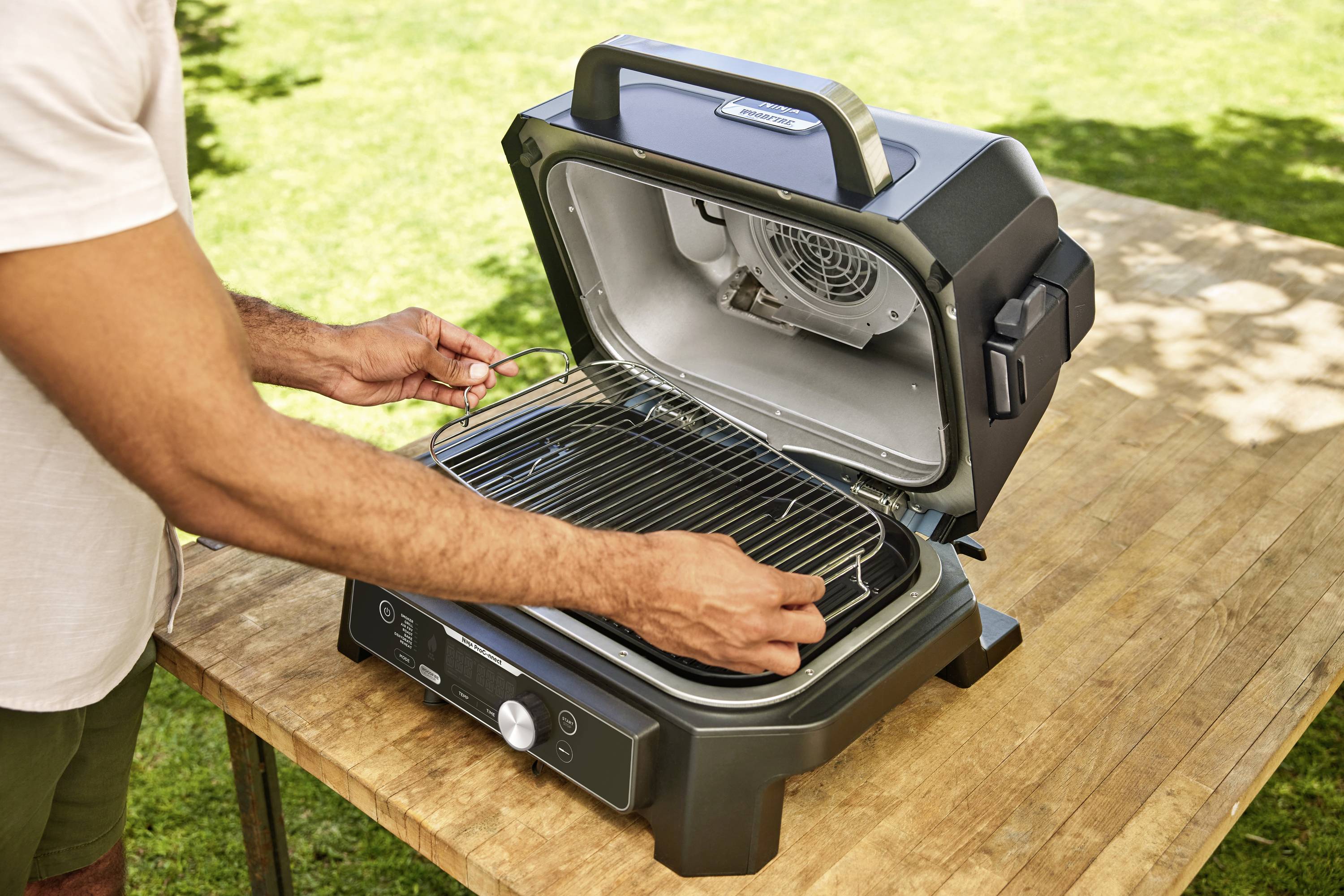 A person places a grill rack into a small electric barbecue outdoors, standing on a wooden table with a sunny garden in the background.